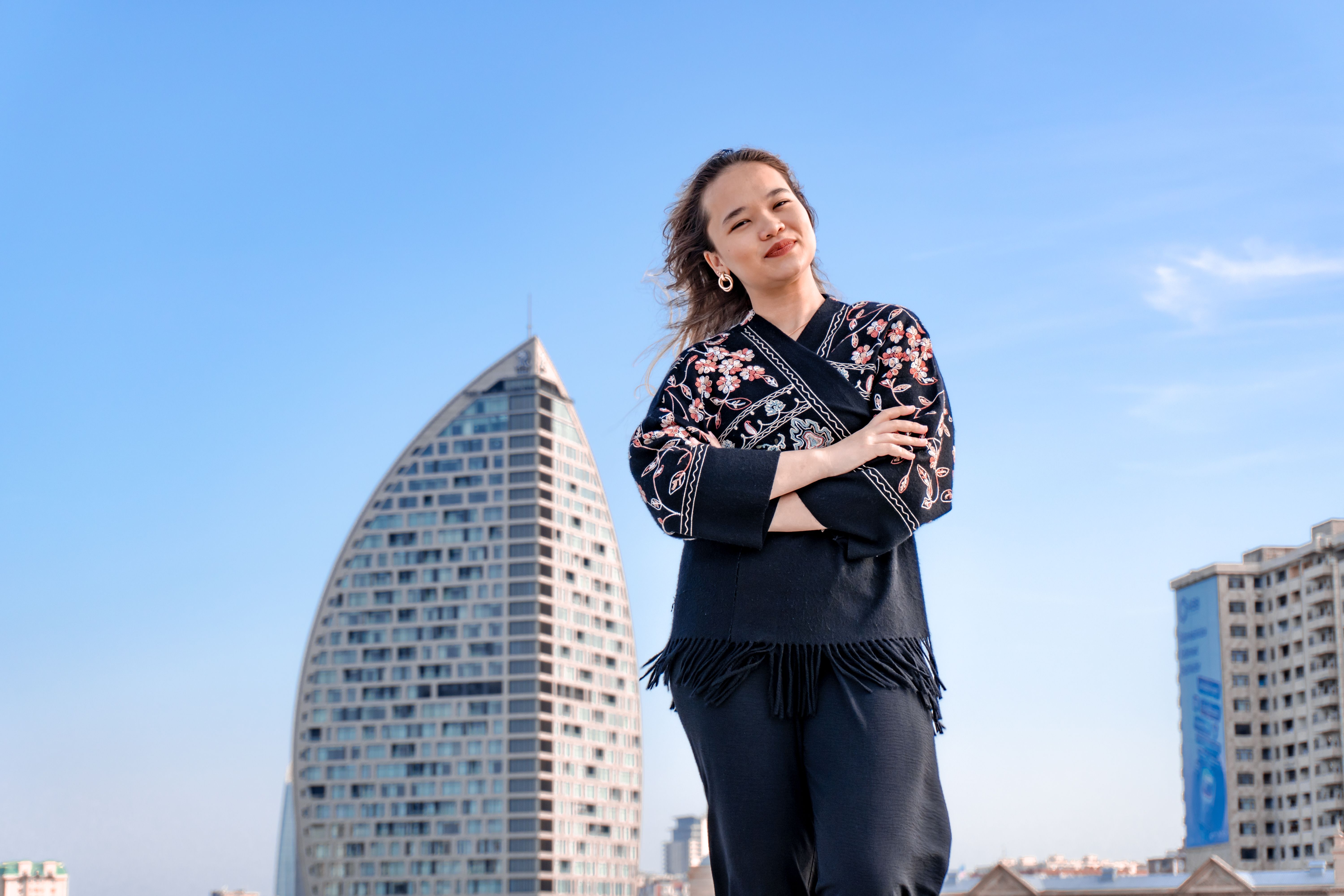 Portrait of a woman in traditional attire standing in front of a modern, sail-shaped building under a clear blue sky.