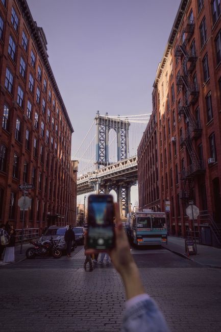 View of Manhattan Bridge framed by red brick buildings in Dumbo, Brooklyn with smartphone focus.