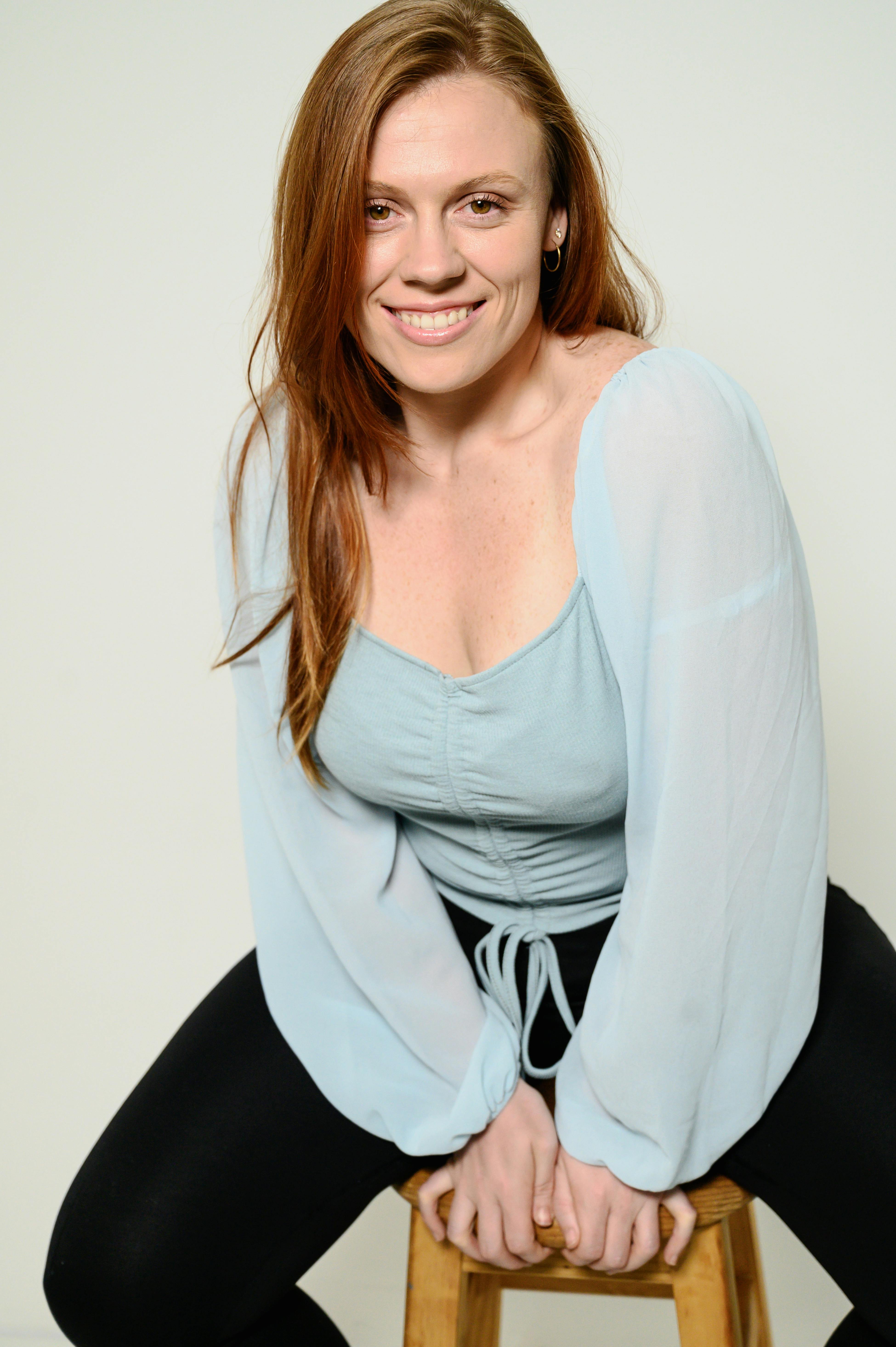 Smiling woman in light blue blouse sitting on a wooden stool. Casual portrait indoors.