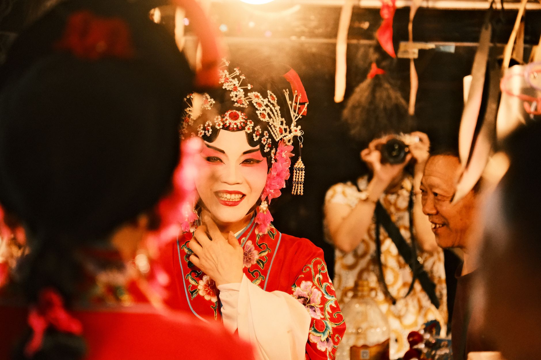 Chinese opera performer in traditional costume and makeup prepares backstage under warm lighting.