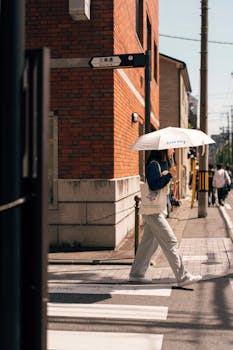 A pedestrian crossing a street in Kyoto with an umbrella on a sunny day.