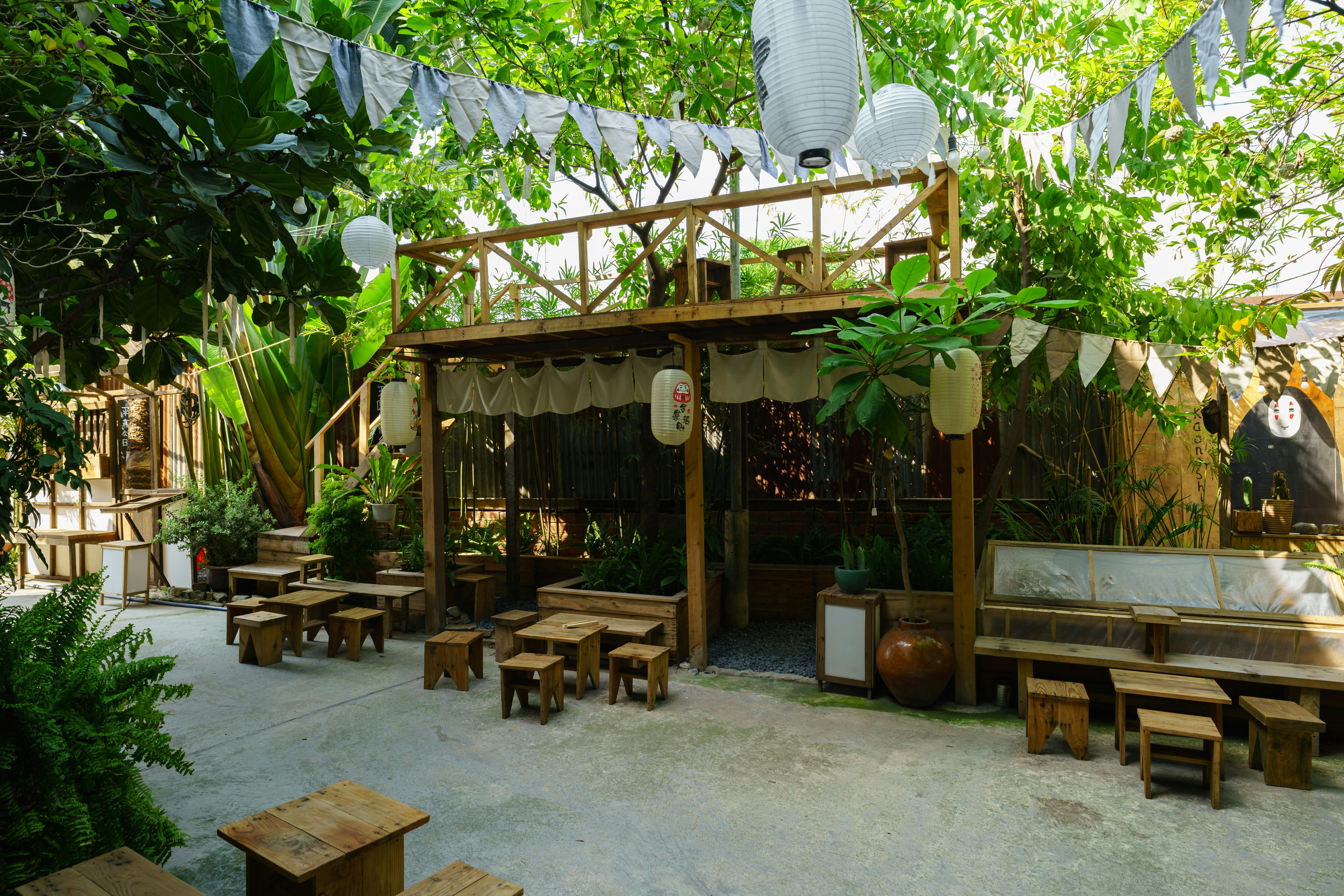 Terrasse de café en plein air avec sièges en bois entourés de verdure luxuriante et de lanternes.