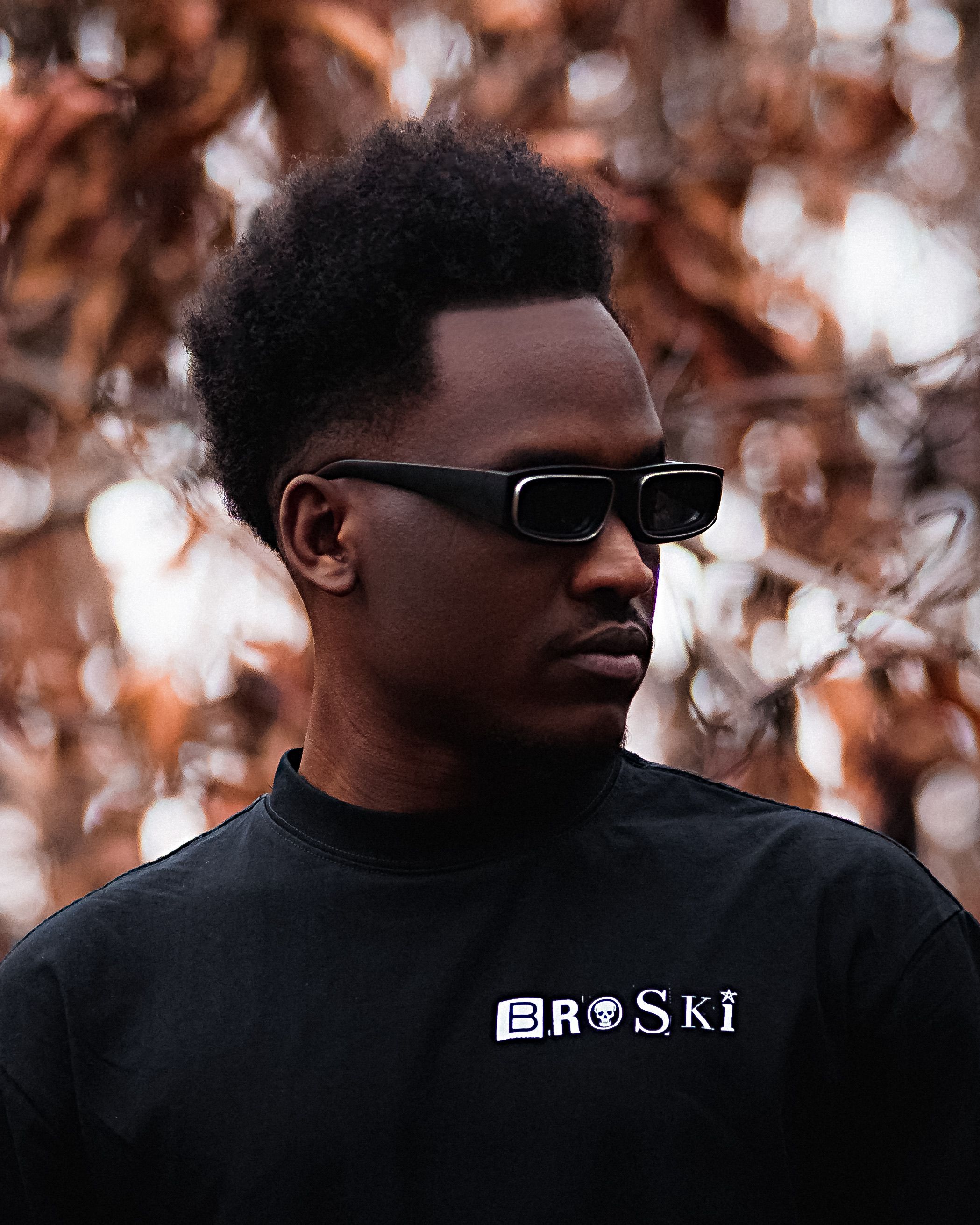 Portrait of a fashionable young man wearing sunglasses and a branded shirt, posing outdoors in autumn.