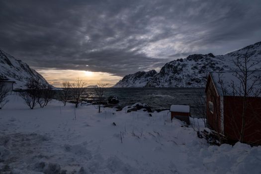 Snow-covered fjord landscape at sunset with dramatic clouds and distant mountains.
