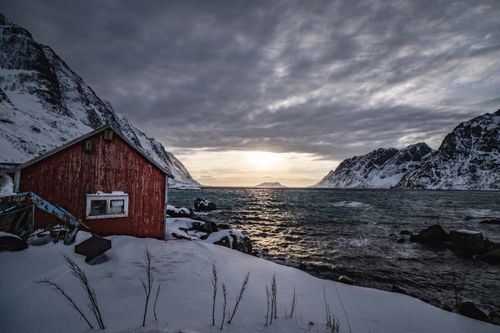 Free A tranquil winter scene in Norway with a red cabin, snow-covered mountains, and a serene lake at sunset. Stock Photo