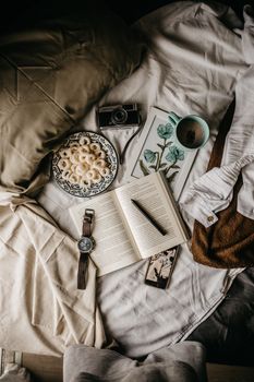 A cozy flat lay featuring a book, coffee, snacks, and a vintage camera on a bed, creating a relaxed atmosphere.