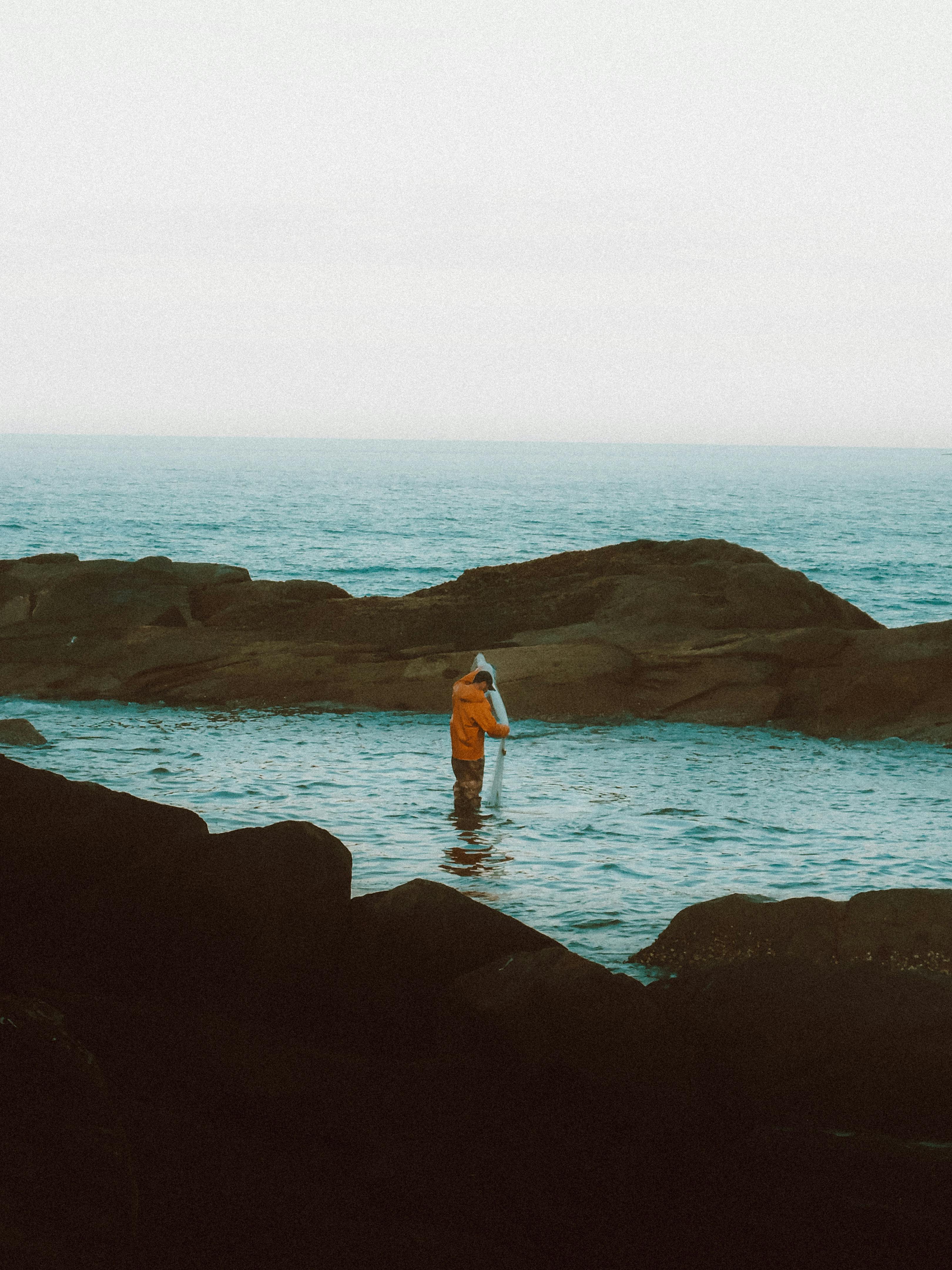A solitary angler in a yellow jacket fishes on a rocky coastline, capturing serene ocean vibes.