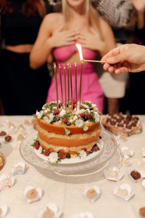 Free Elegant birthday cake with candles being lit, surrounded by guests celebrating indoors. Stock Photo
