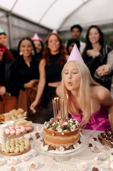 Woman blowing birthday candles surrounded by cheerful friends and delicious desserts.