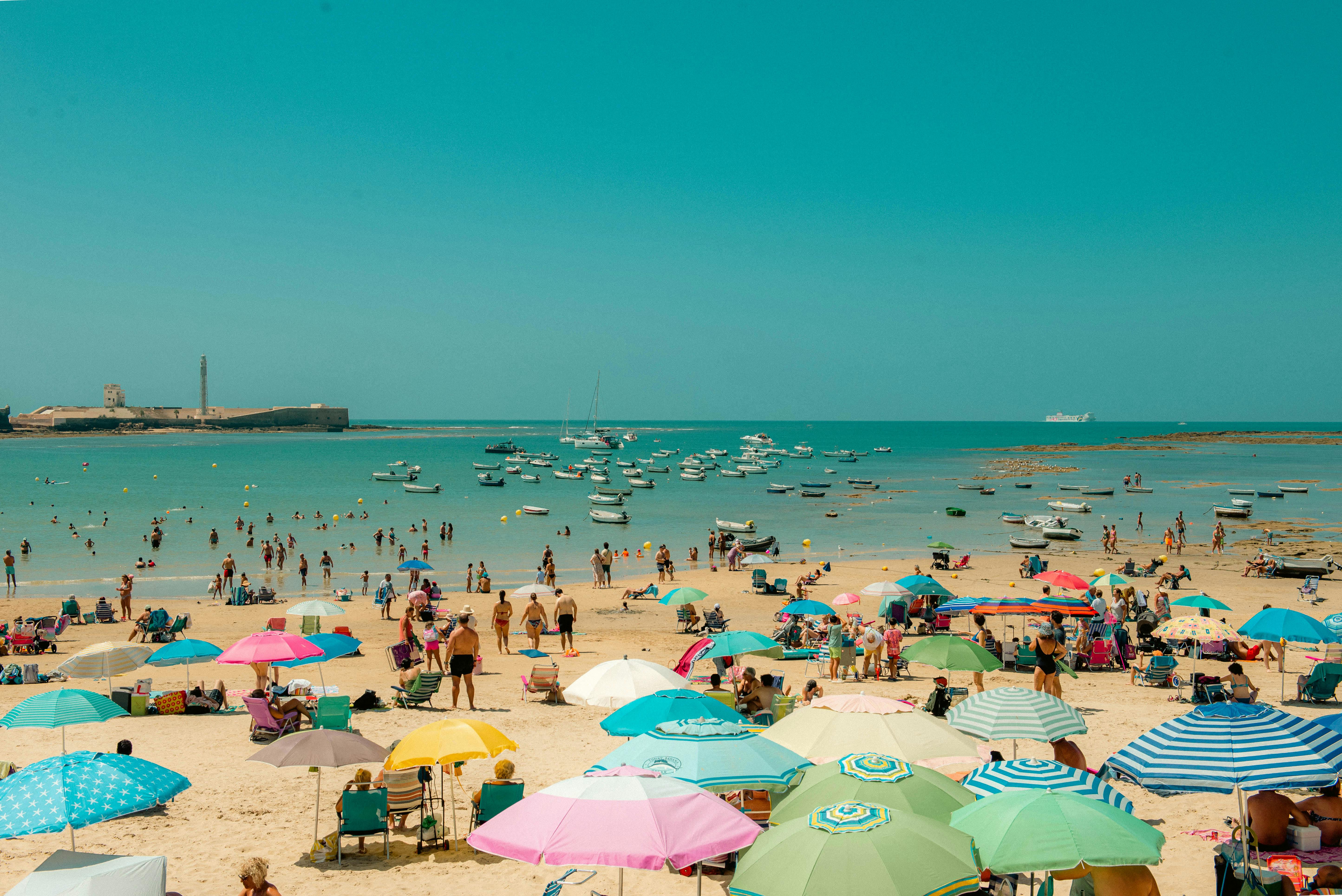 Crowded beach scene with colorful umbrellas, people swimming, and boats anchored near the shore under a clear sky.