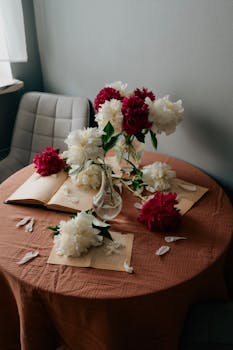 Beautiful red and white peony bouquet in a glass vase on a round table with books, creating a cozy ambiance.