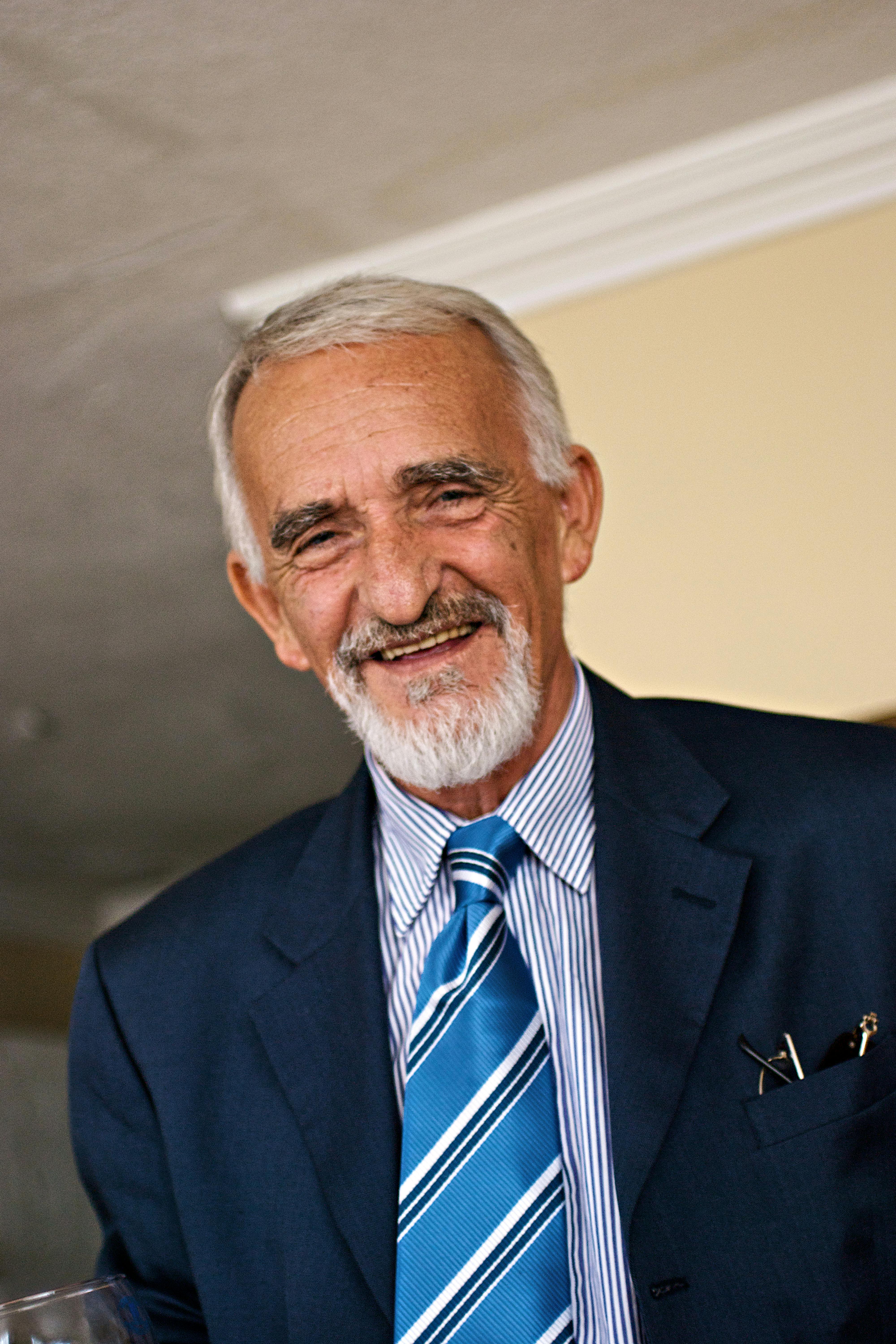 Senior man in a blue suit smiles warmly indoors, exuding charm and wisdom.
