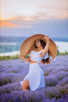 A woman in a white dress poses with a large hat in a vibrant lavender field during sunset.