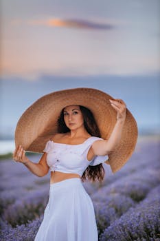 Elegant woman in white dress and large hat in a vibrant lavender field during sunset.