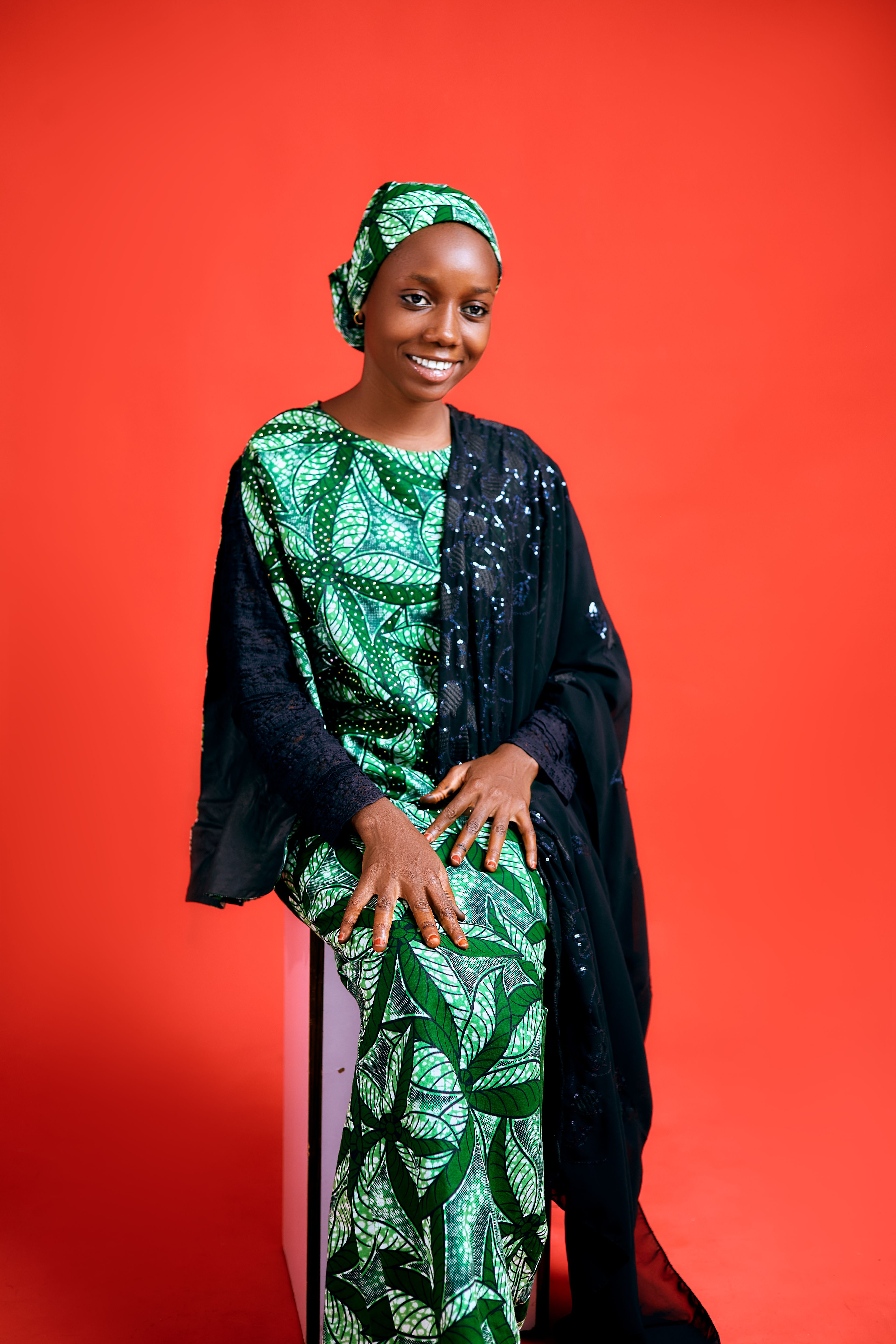 Portrait of a smiling Nigerian woman in vibrant traditional attire, set against a bold red background.