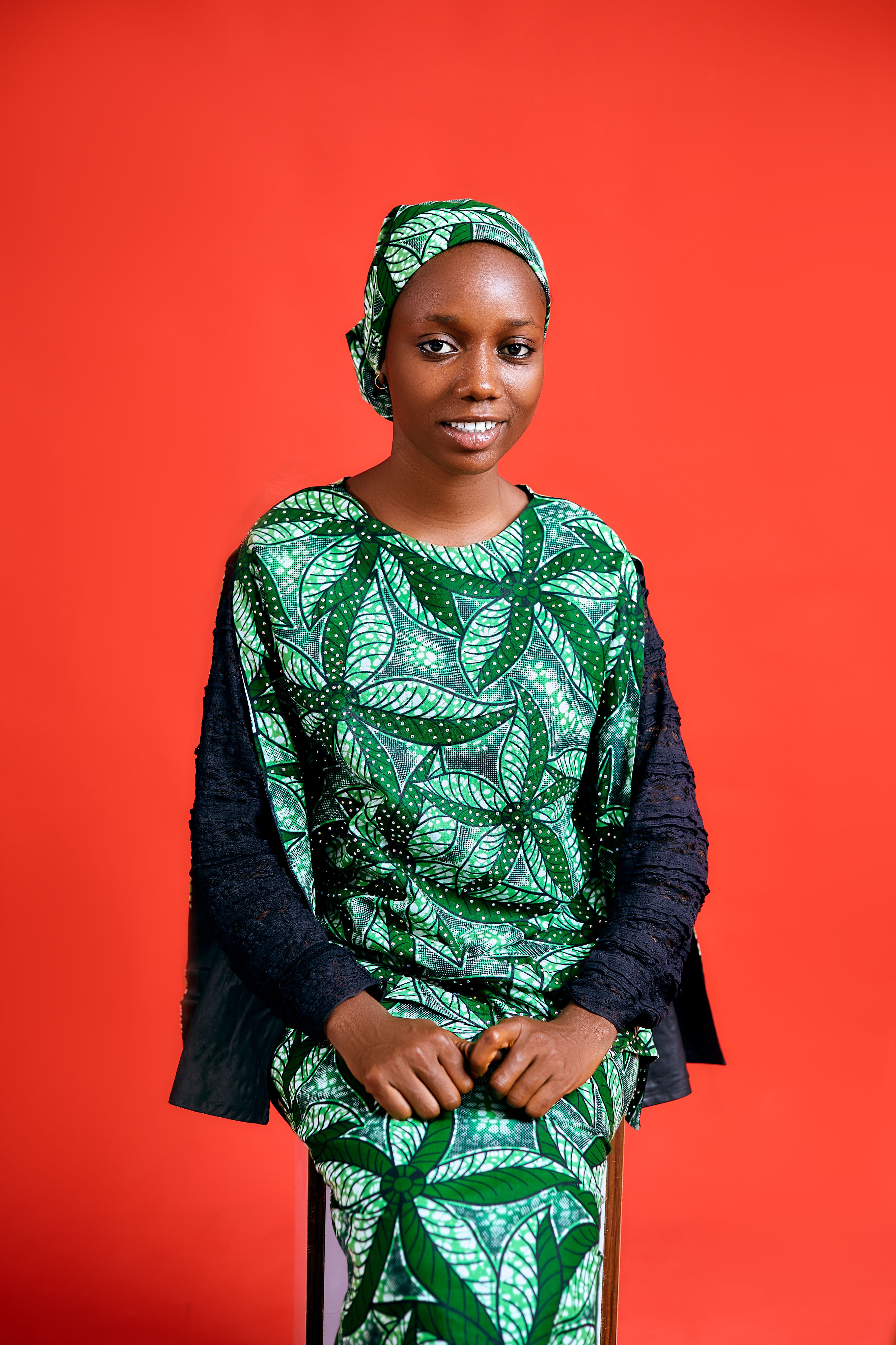 A woman in colorful African attire sits against an orange background, smiling.