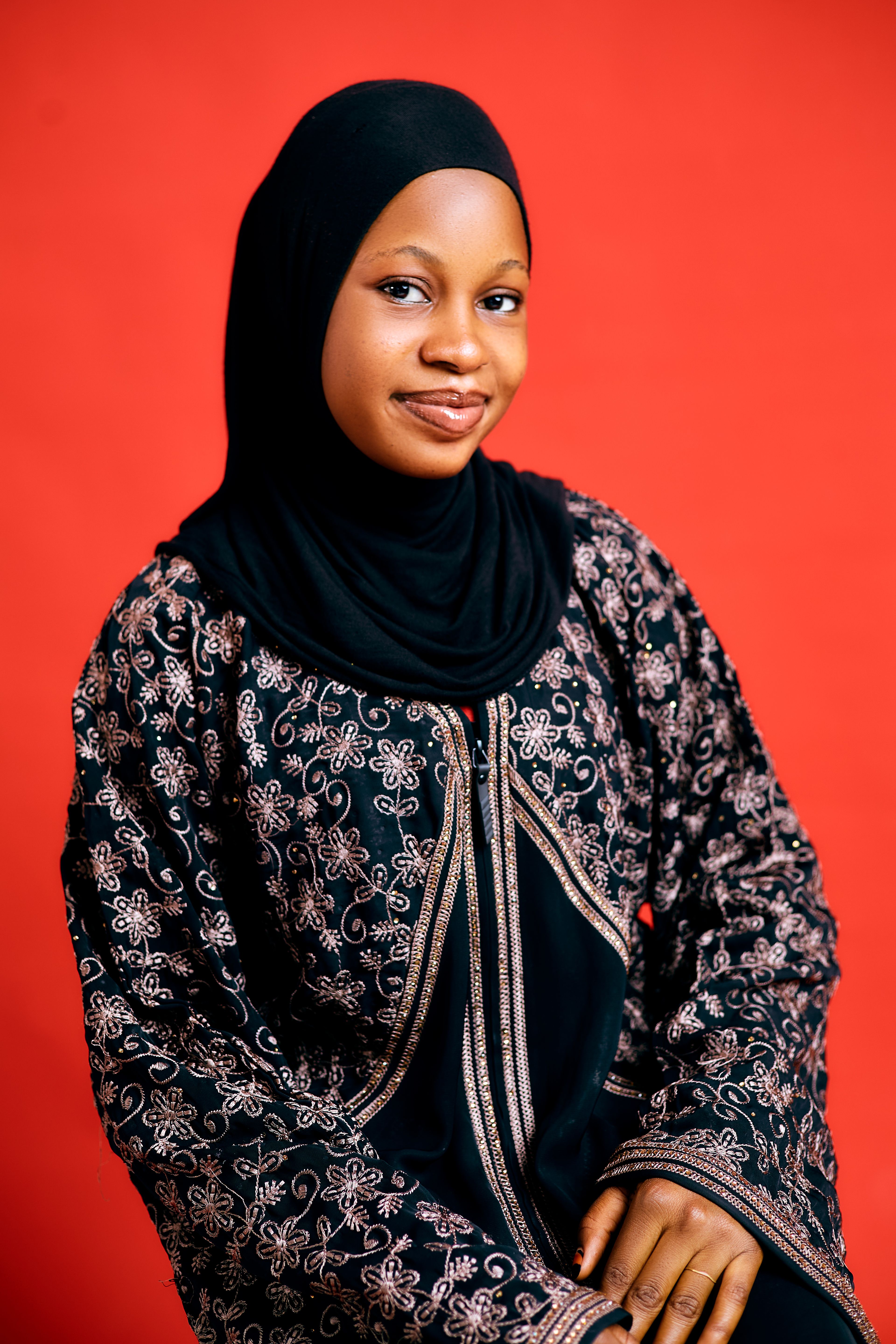 A woman wearing a black hijab and intricate embroidered dress against a red background.