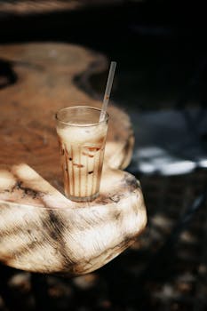 A refreshing iced coffee in a glass with straw on a rustic wooden table under dappled sunlight.