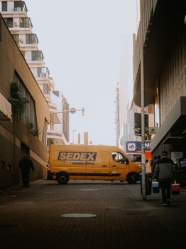 Yellow delivery van in a city street, alongside modern architecture and pedestrians.