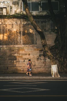 A person walking by a weathered stone wall with trees in an urban setting.