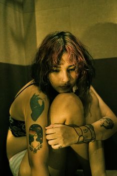 A contemplative woman with tattoos sits alone in lingerie, evoking a moody and introspective atmosphere.