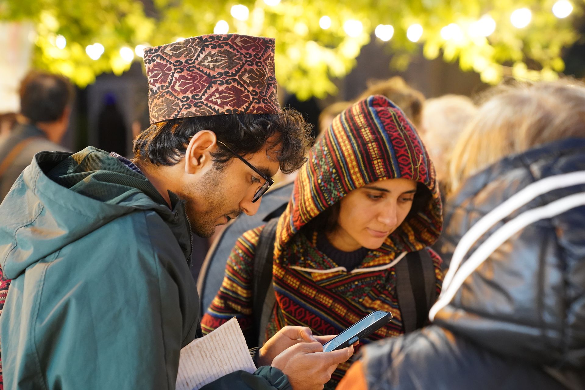 Two adults engaged with a smartphone at an outdoor social event under string lights.