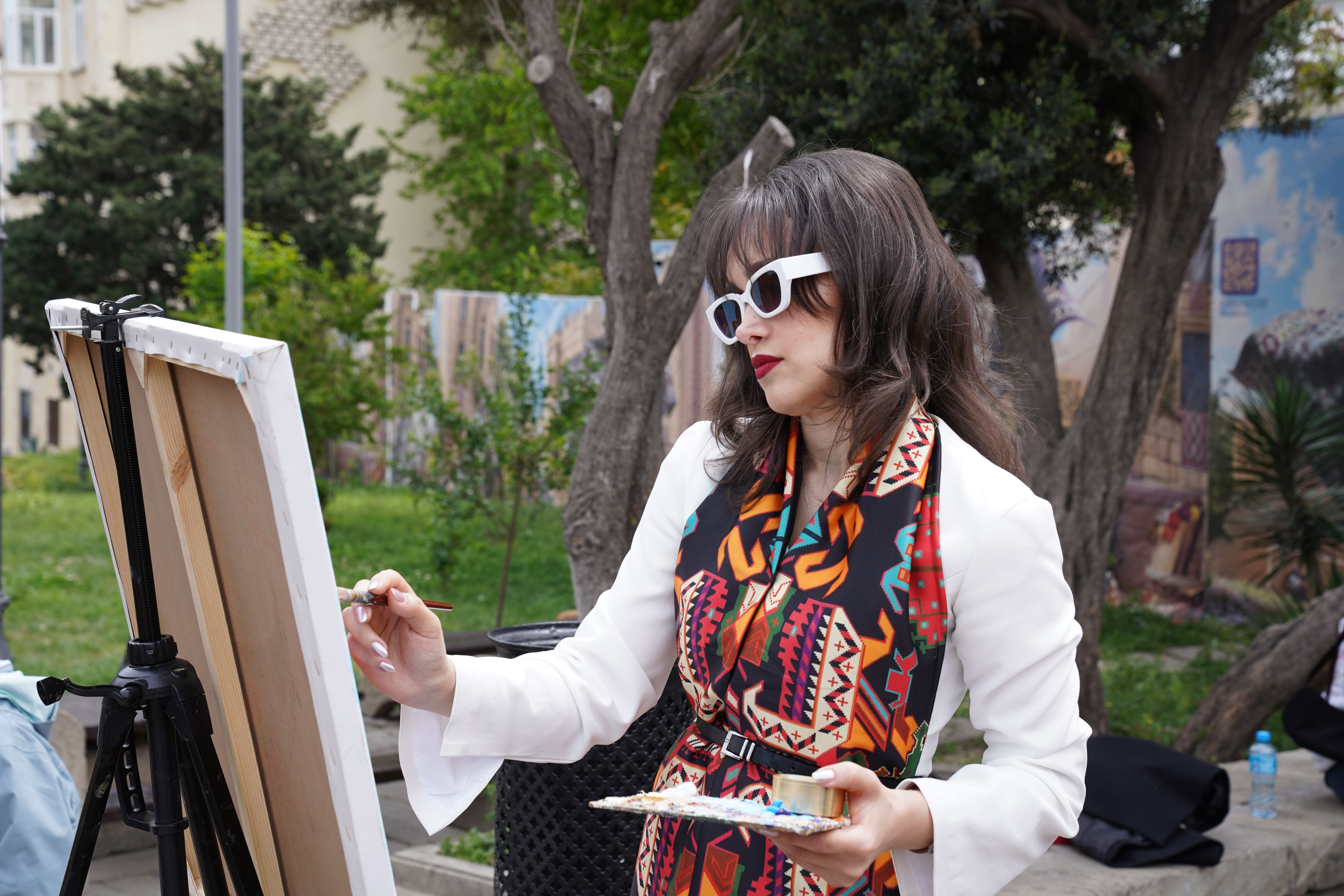 Stylish woman painting outdoors with vibrant scarf and sunglasses amidst greenery.