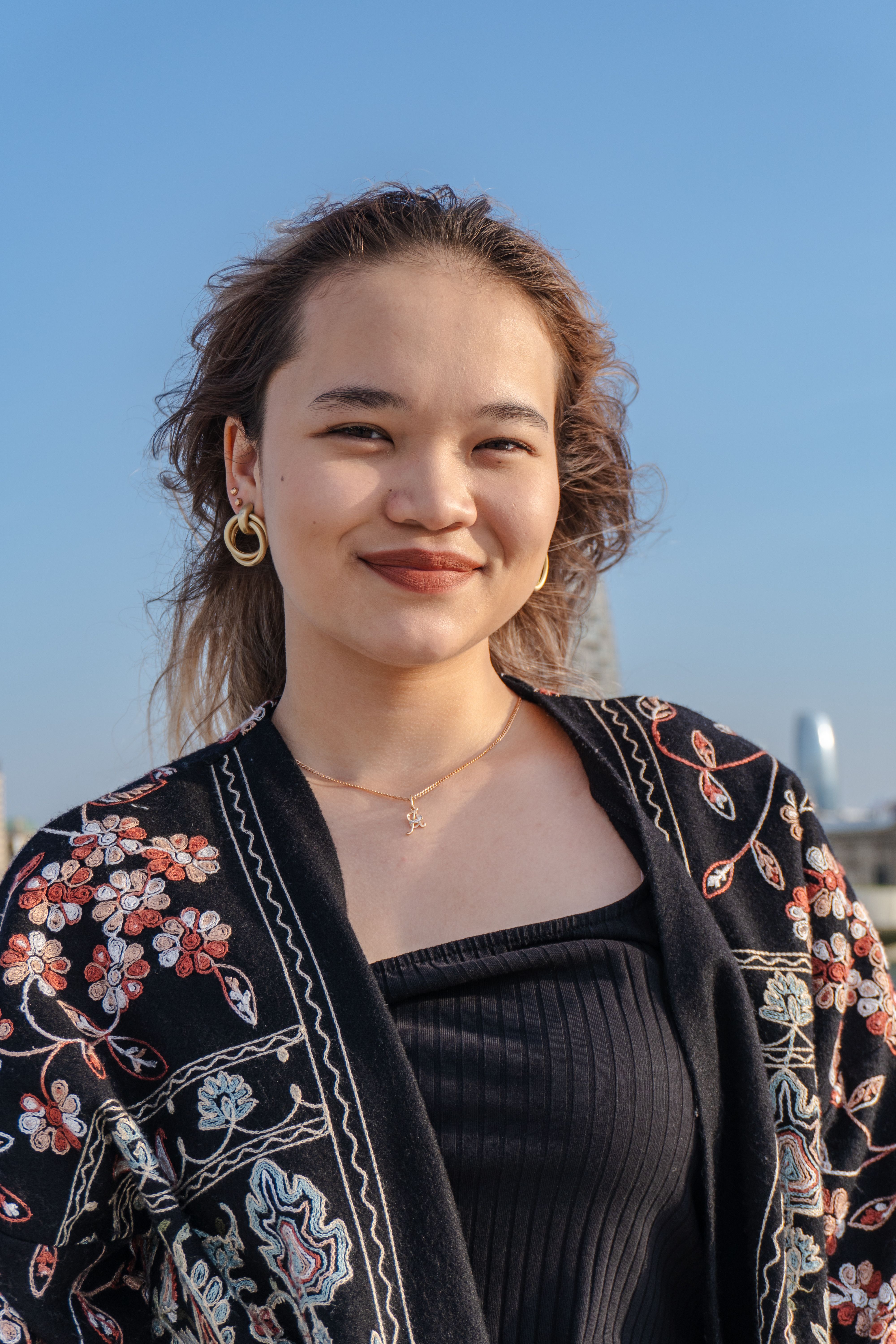 A young woman smiling outdoors under a clear blue sky, showcasing earrings and a floral jacket.