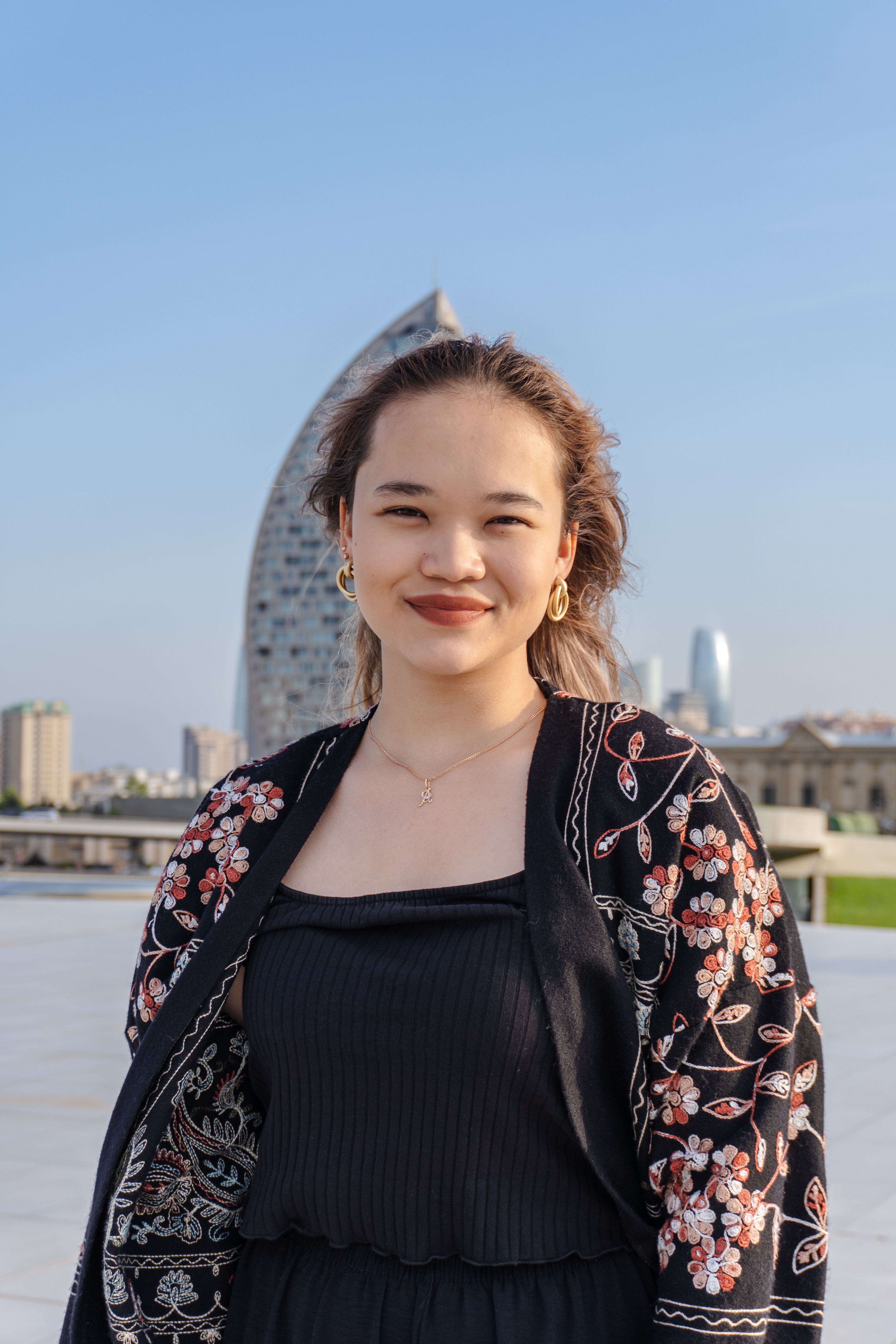Casual portrait of a young woman smiling outdoors with the Baku skyline in the background on a sunny day.