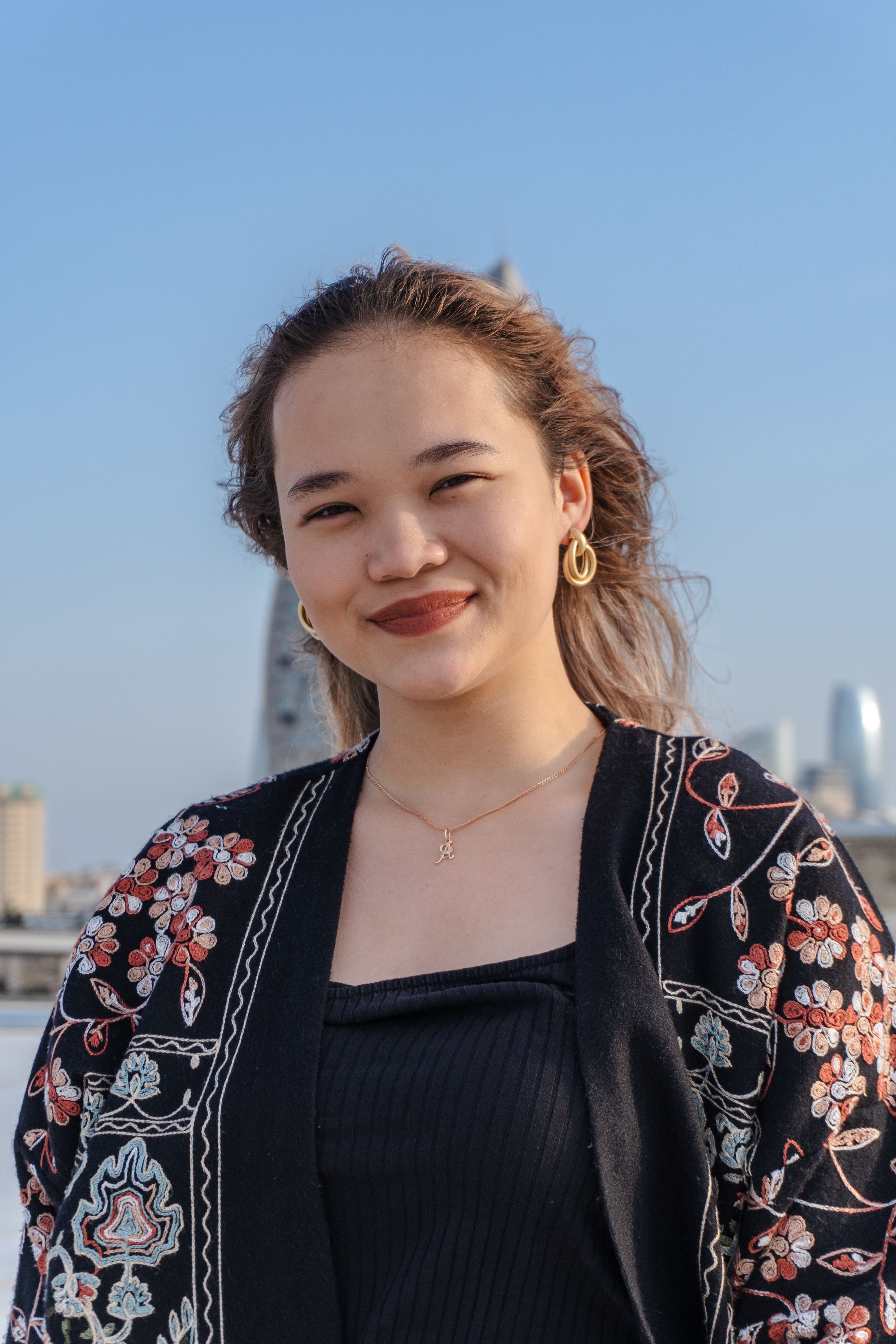 Portrait of a young woman smiling under a clear blue sky with an urban cityscape in the background.
