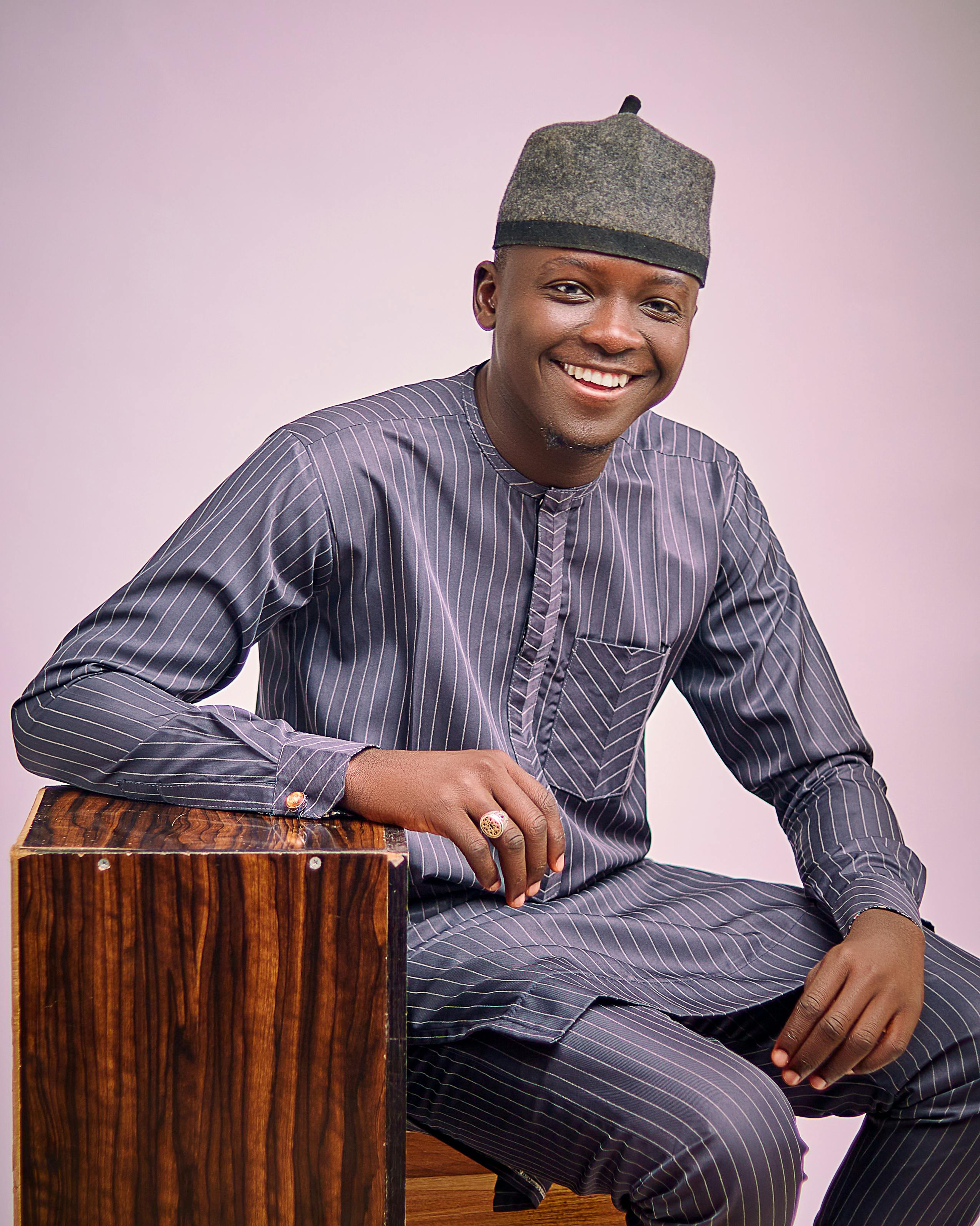 Young man in traditional attire smiling while seated on a wooden box against a neutral background.