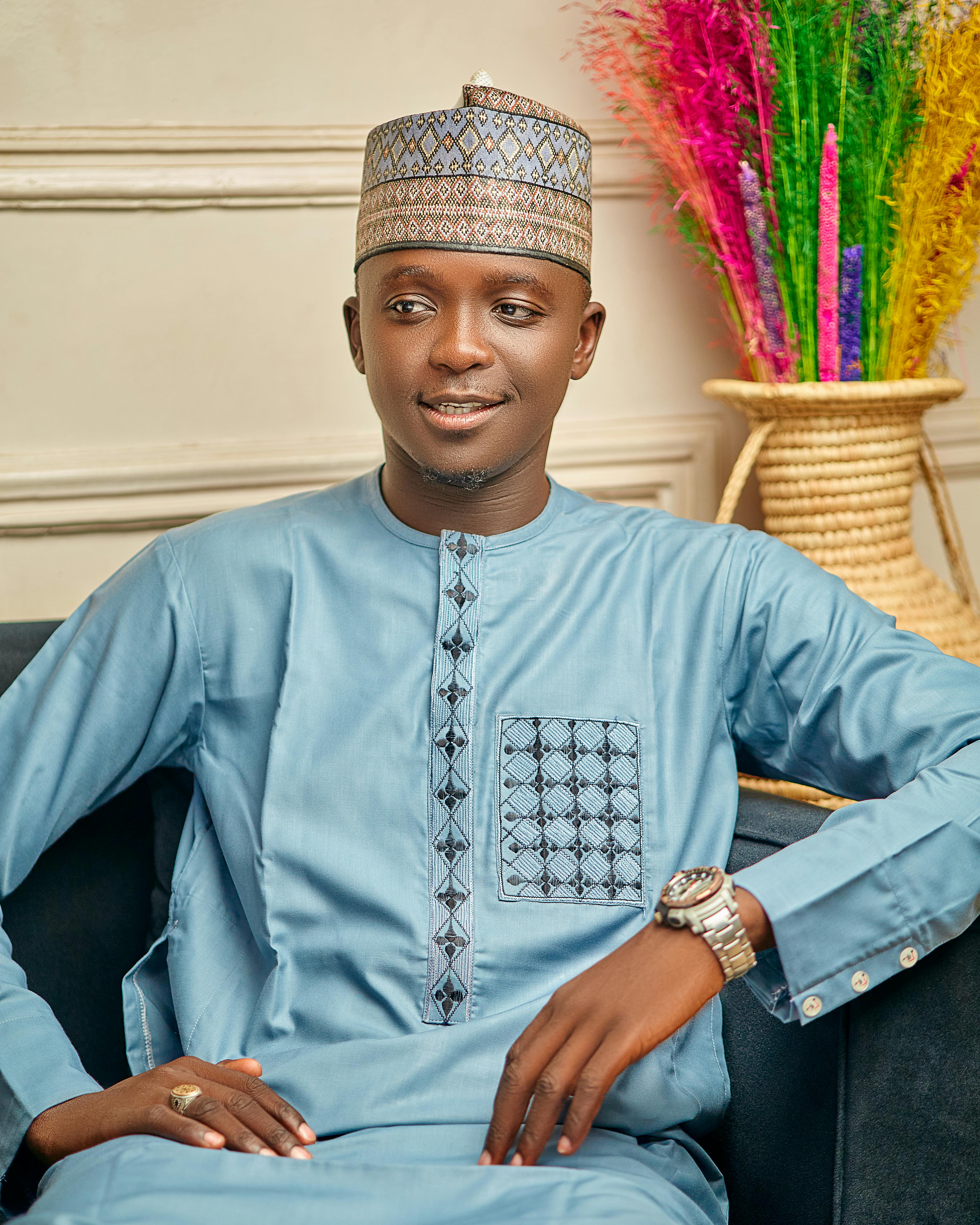 Man wearing light blue traditional attire seated comfortably indoors with colorful decor.
