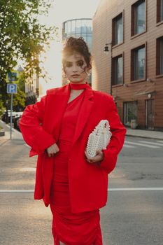 Stylish woman in red dress and blazer holding a white purse in urban setting during sunset.