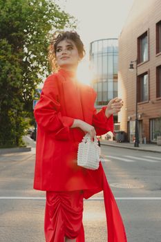 Stylish woman wearing a red outfit and holding a white purse on a sunlit city street.
