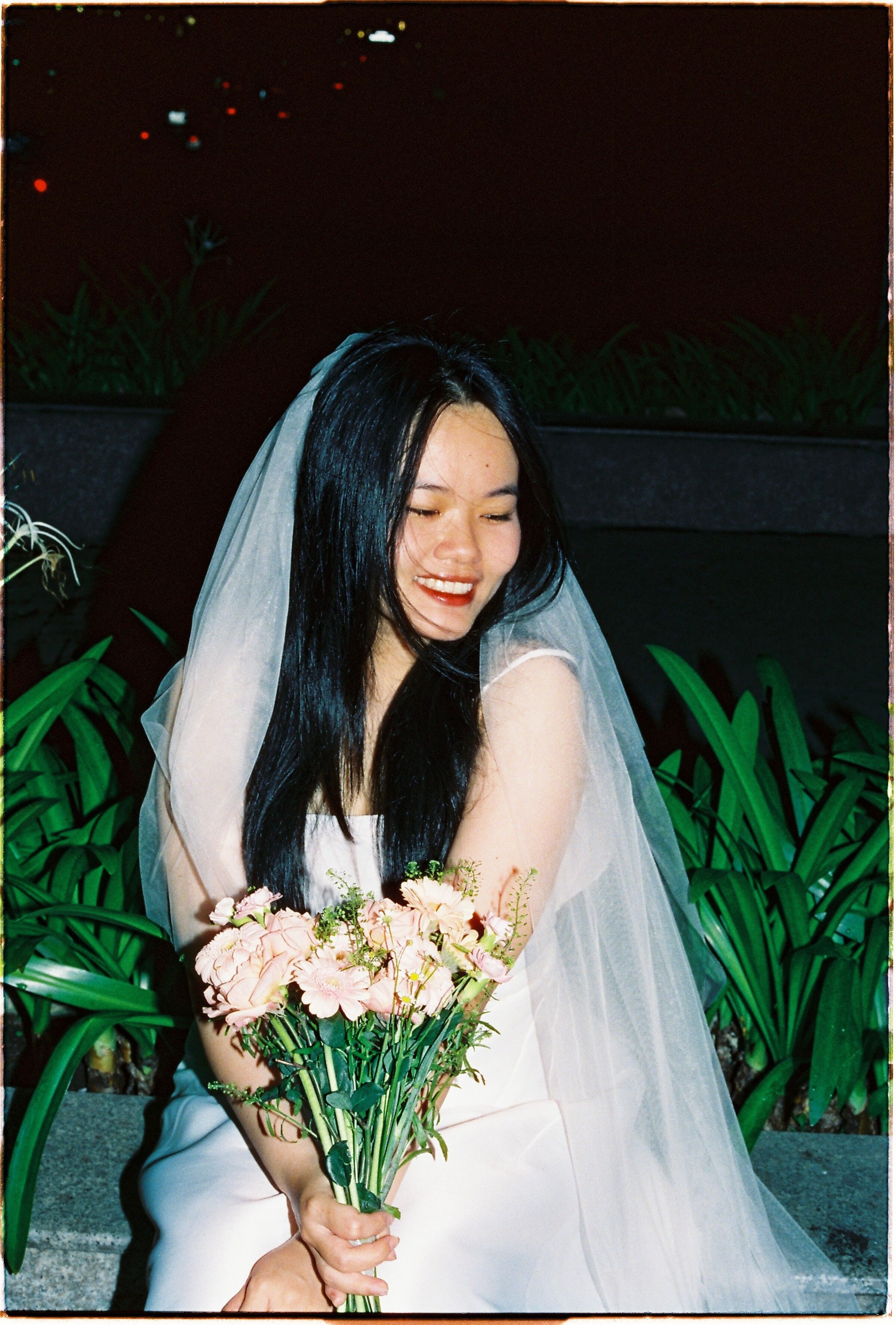 Smiling bride in a veil holding pink flowers outdoors at night.