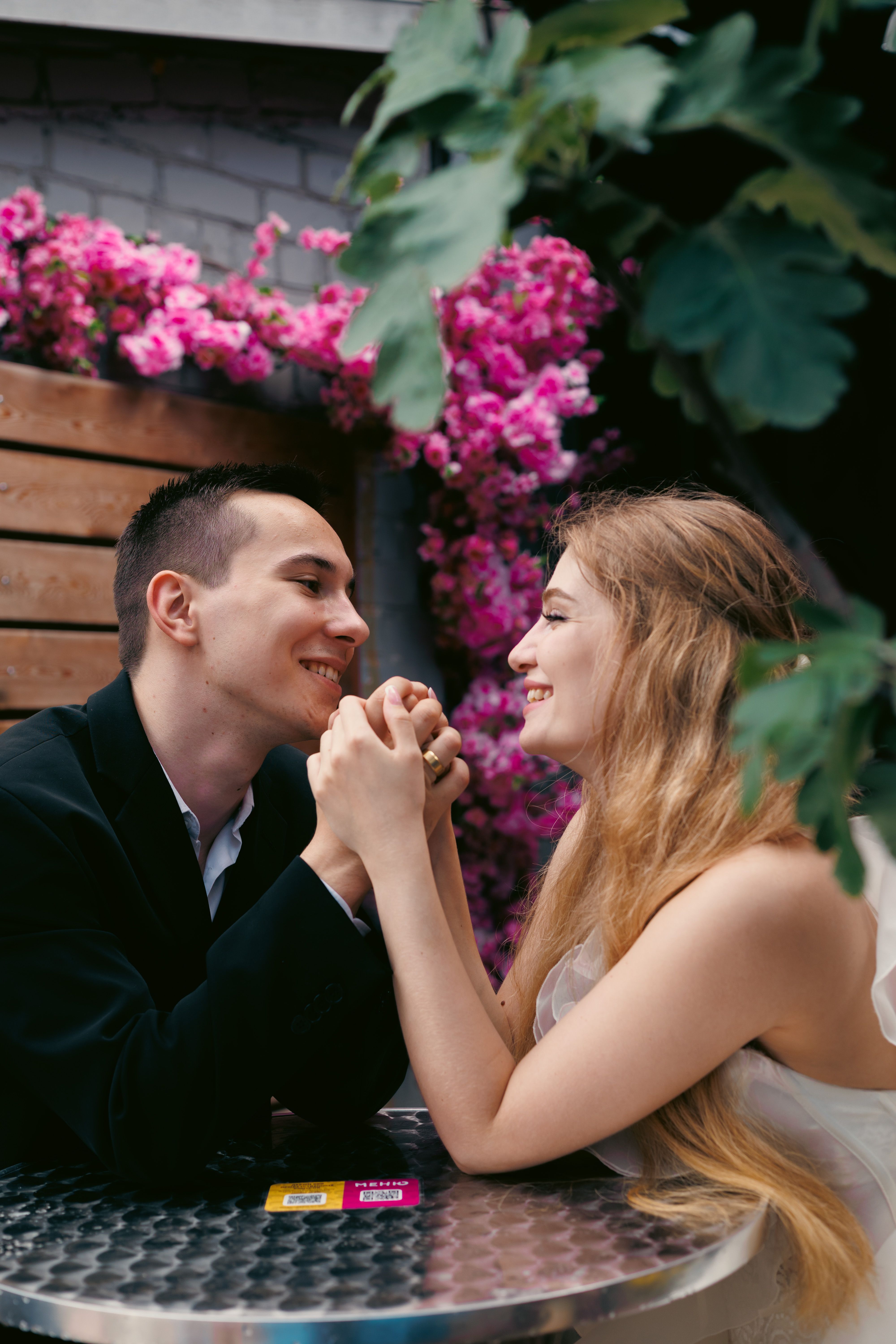 Young couple smiling across a cafe table surrounded by pink flowers, enjoying an outdoor moment.
