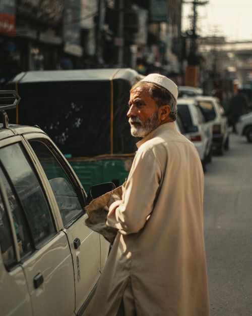 Free An older man in traditional attire stands beside a car in a bustling street scene, showcasing urban life. Stock Photo