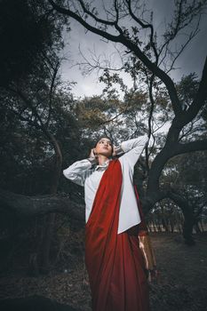 A striking portrait of a woman in a red saree amidst dark, dramatic woodland scenery.