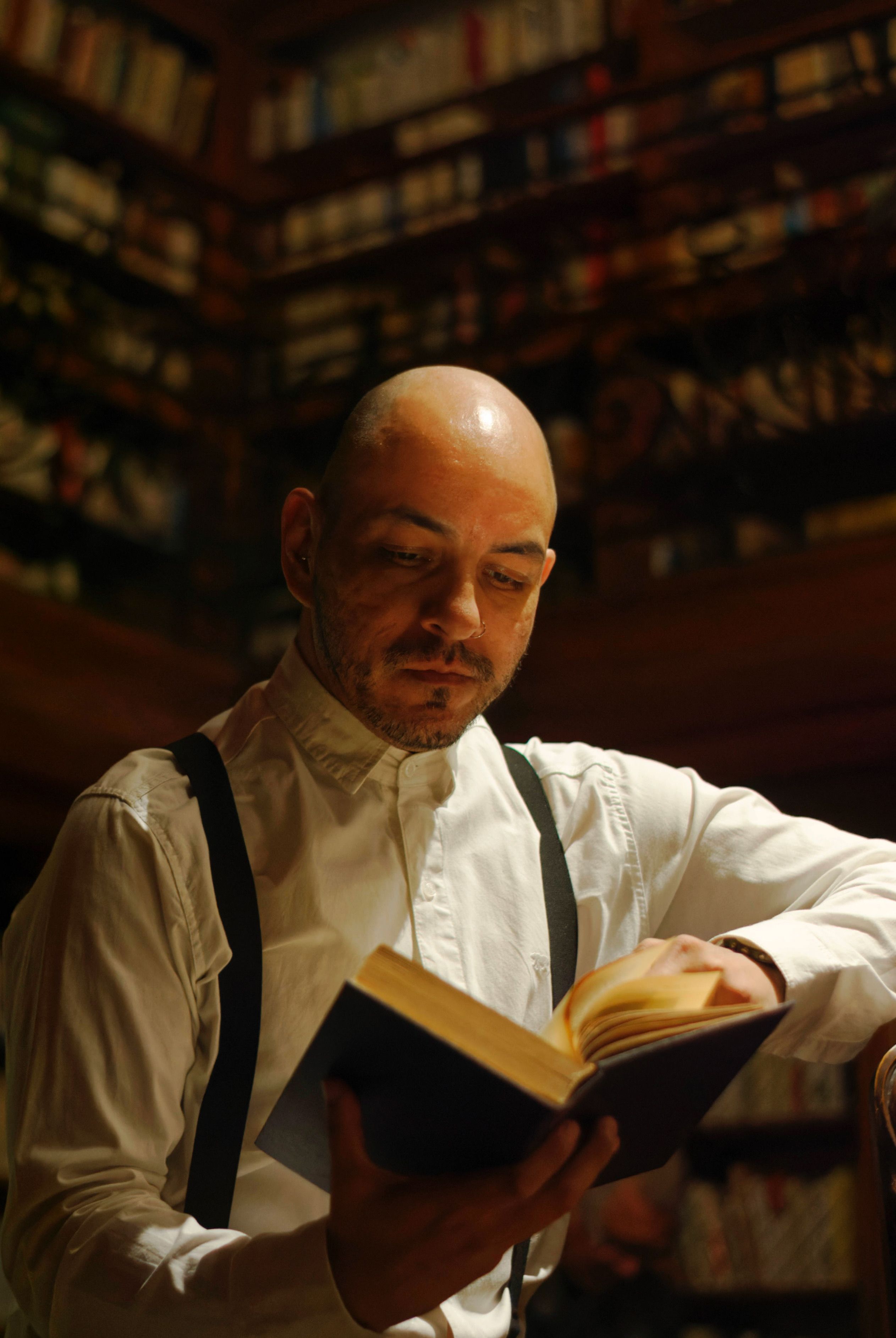 Bald man in white shirt reading a book in a cozy, dimly lit library.