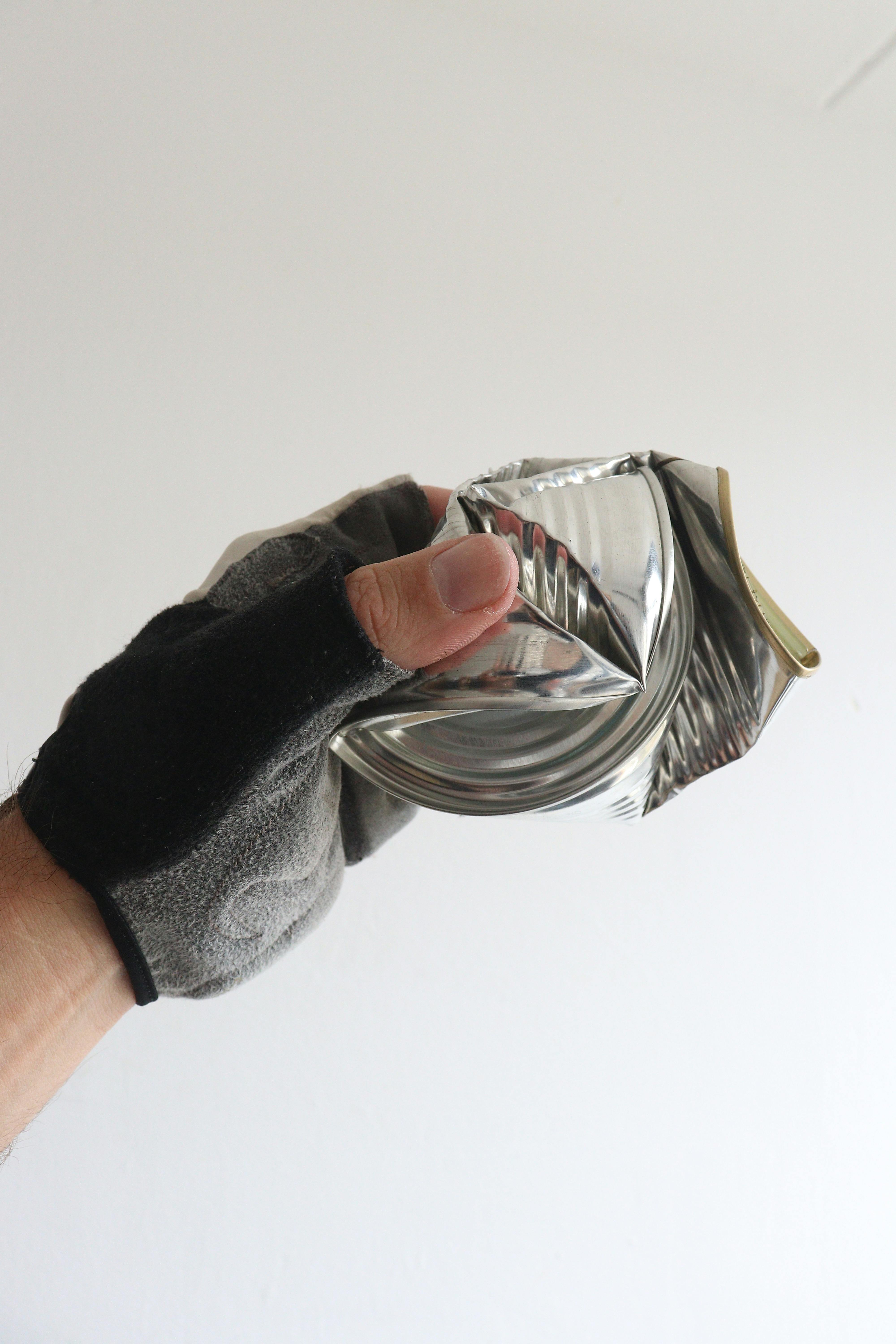 Close-up of a hand in a glove crushing an aluminum can against a plain background.