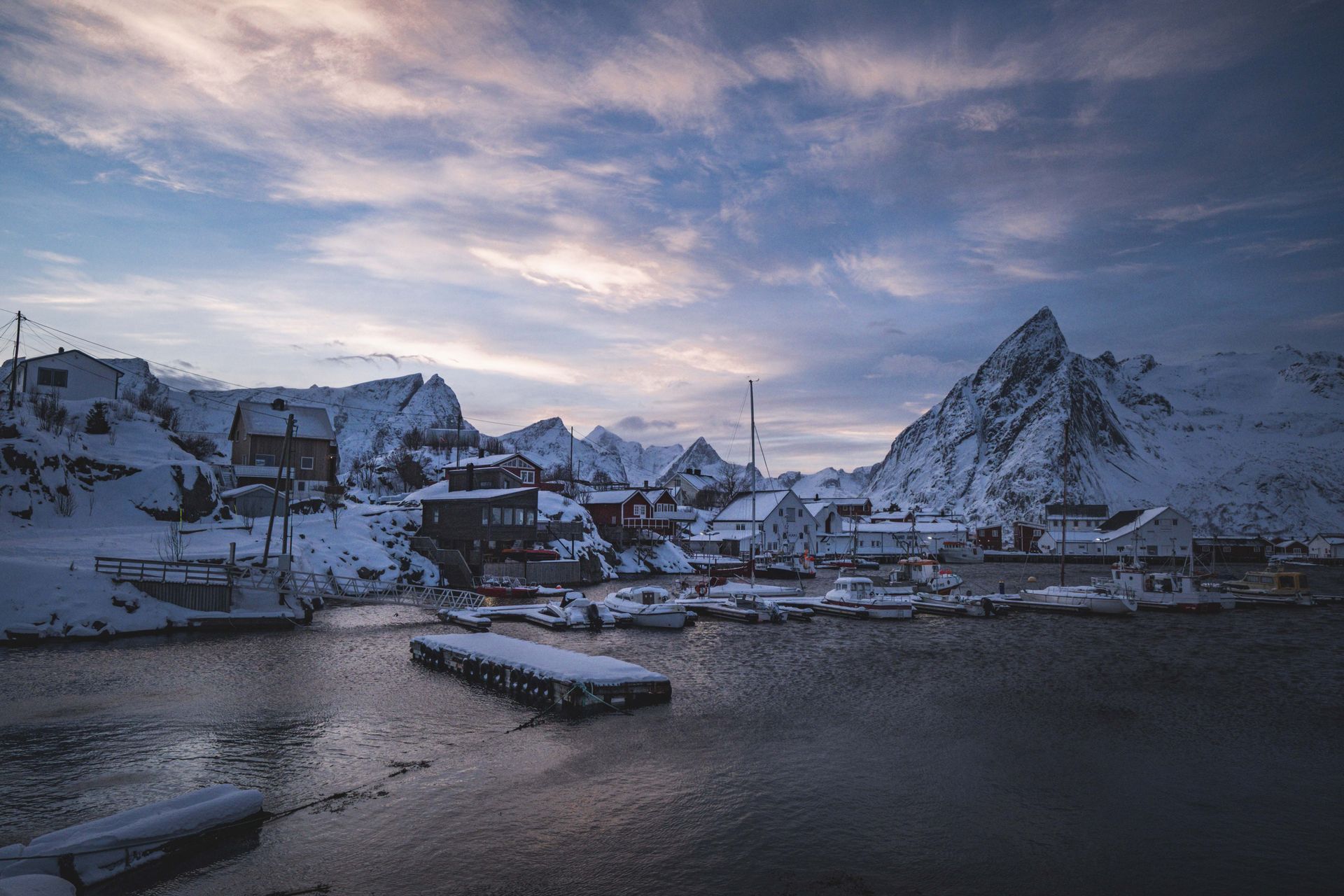 Peaceful winter harbor in Reine, Norway with snowy mountains and boats under a dramatic sky.