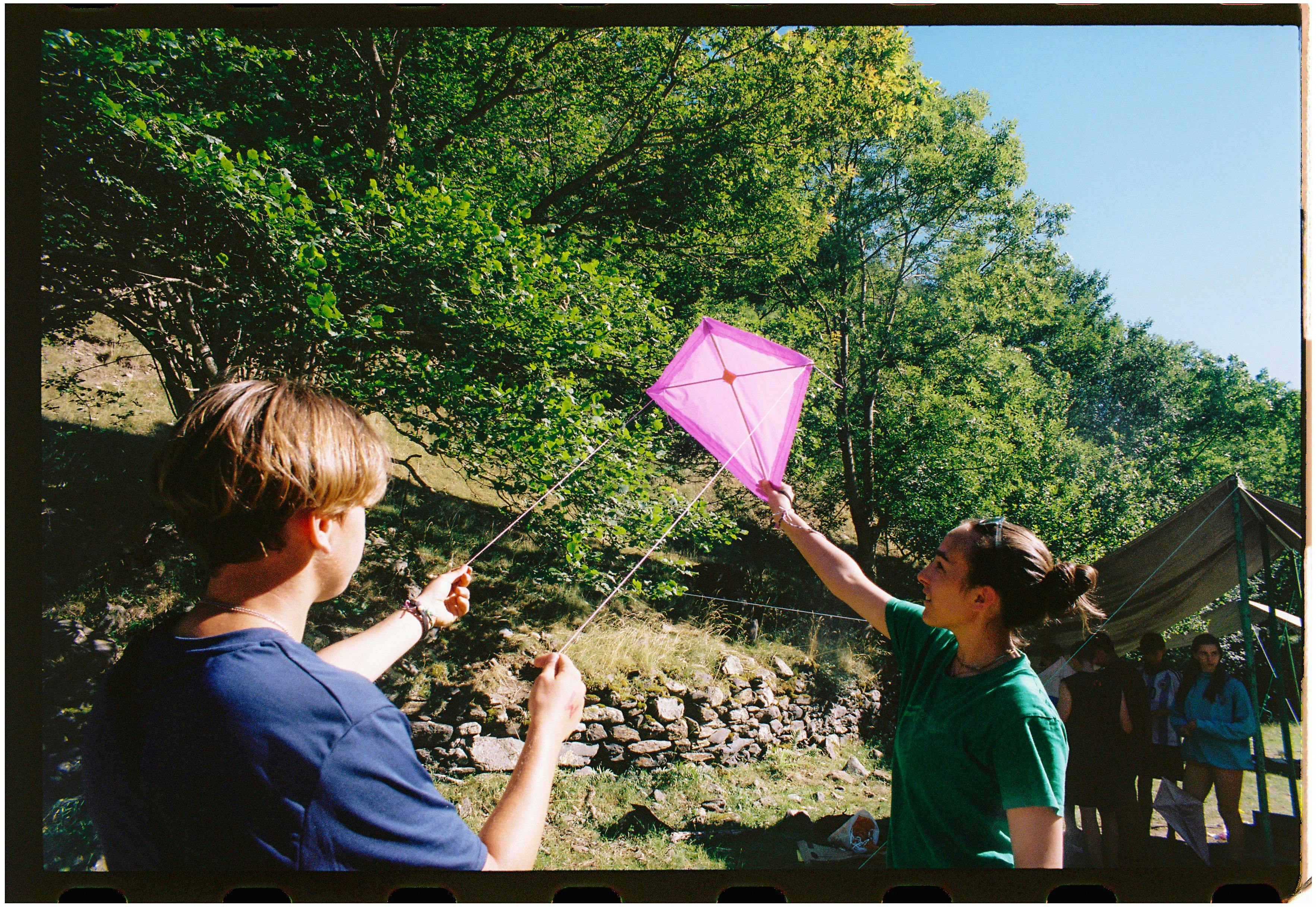 Two young adults fly a pink kite in a lush, sunny outdoor setting, enjoying a leisure day.