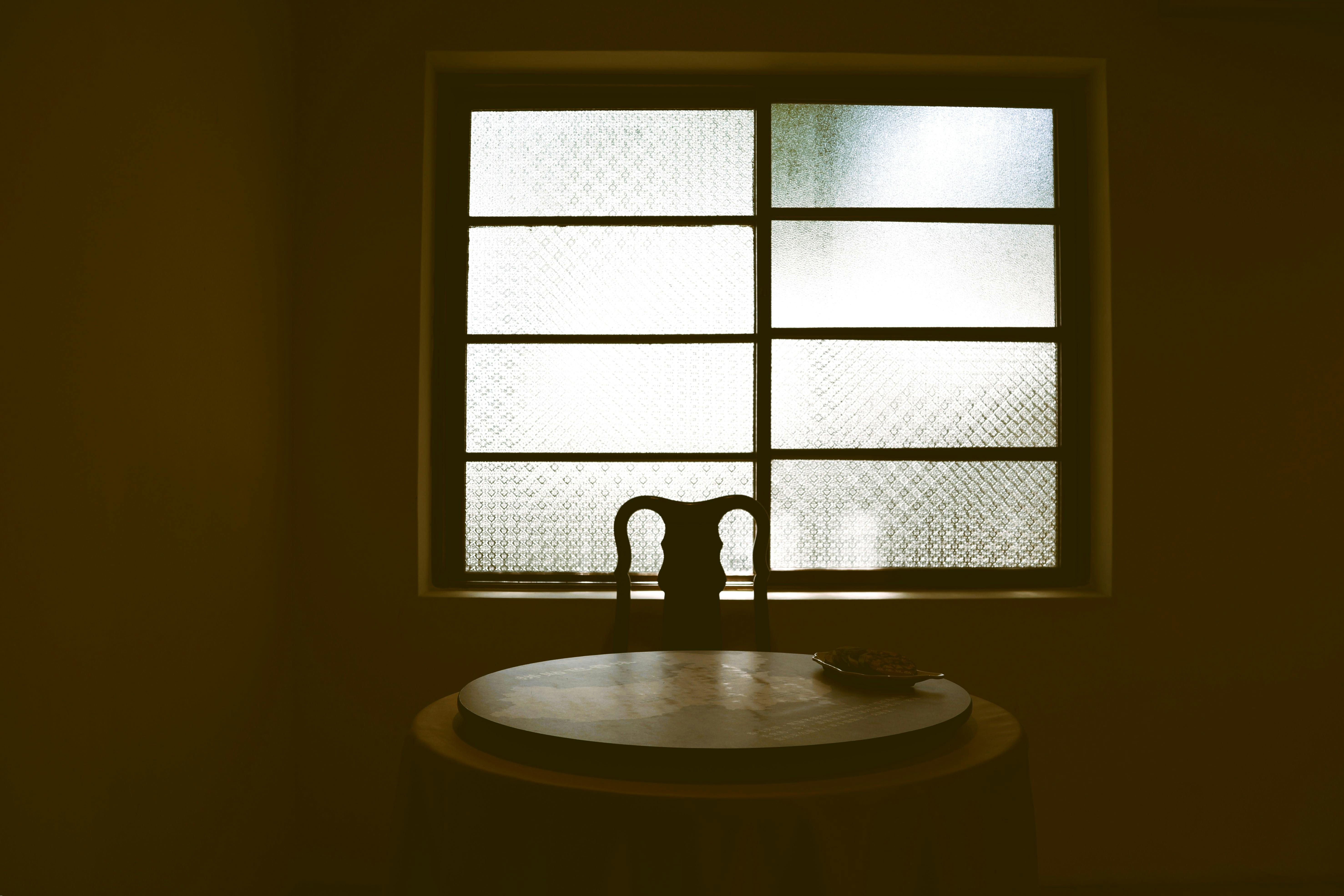 A vintage setup with a chair and round table silhouetted against a textured window.