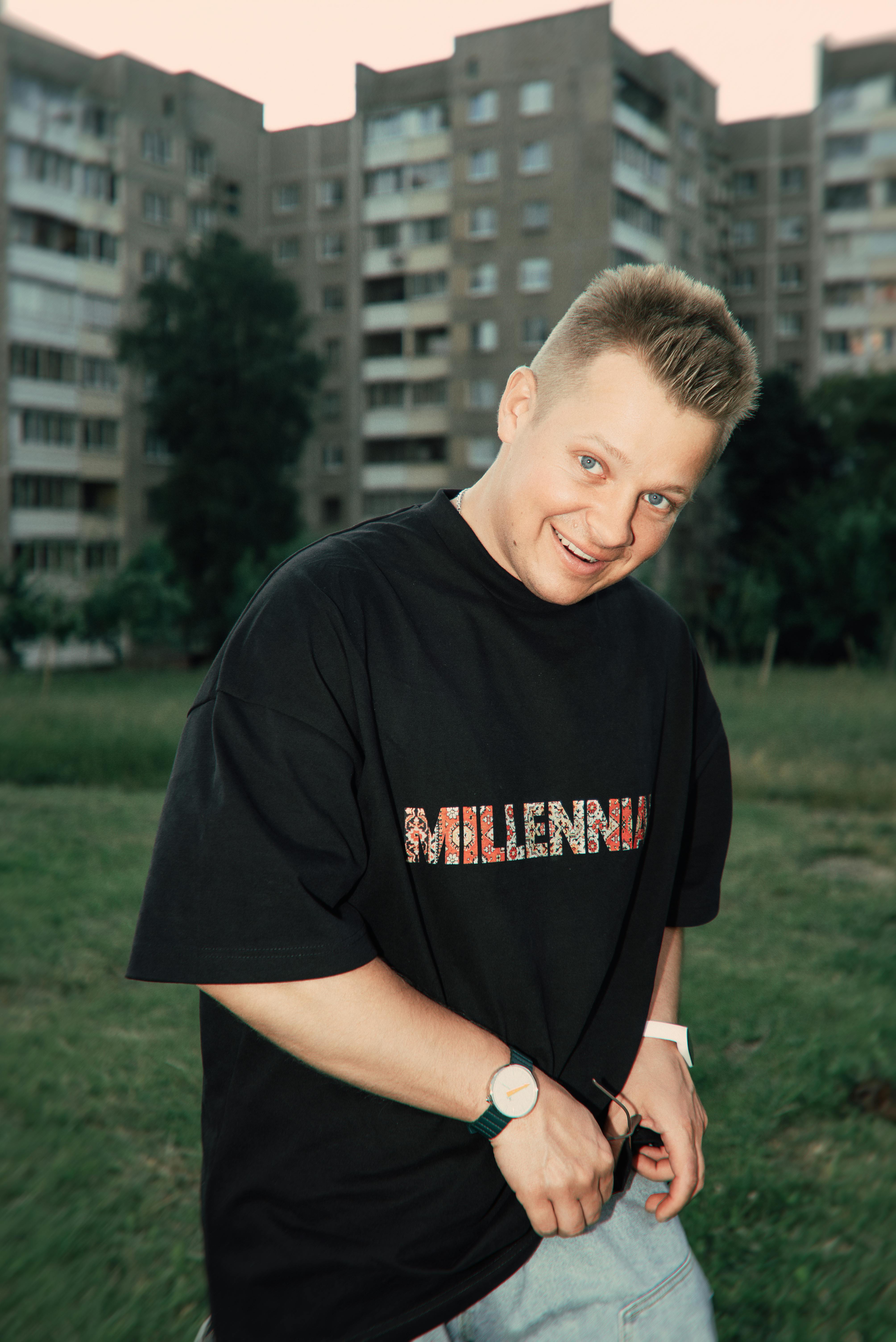 Casual young man smiling outdoors with apartment buildings in the background, wearing a Millennium shirt.