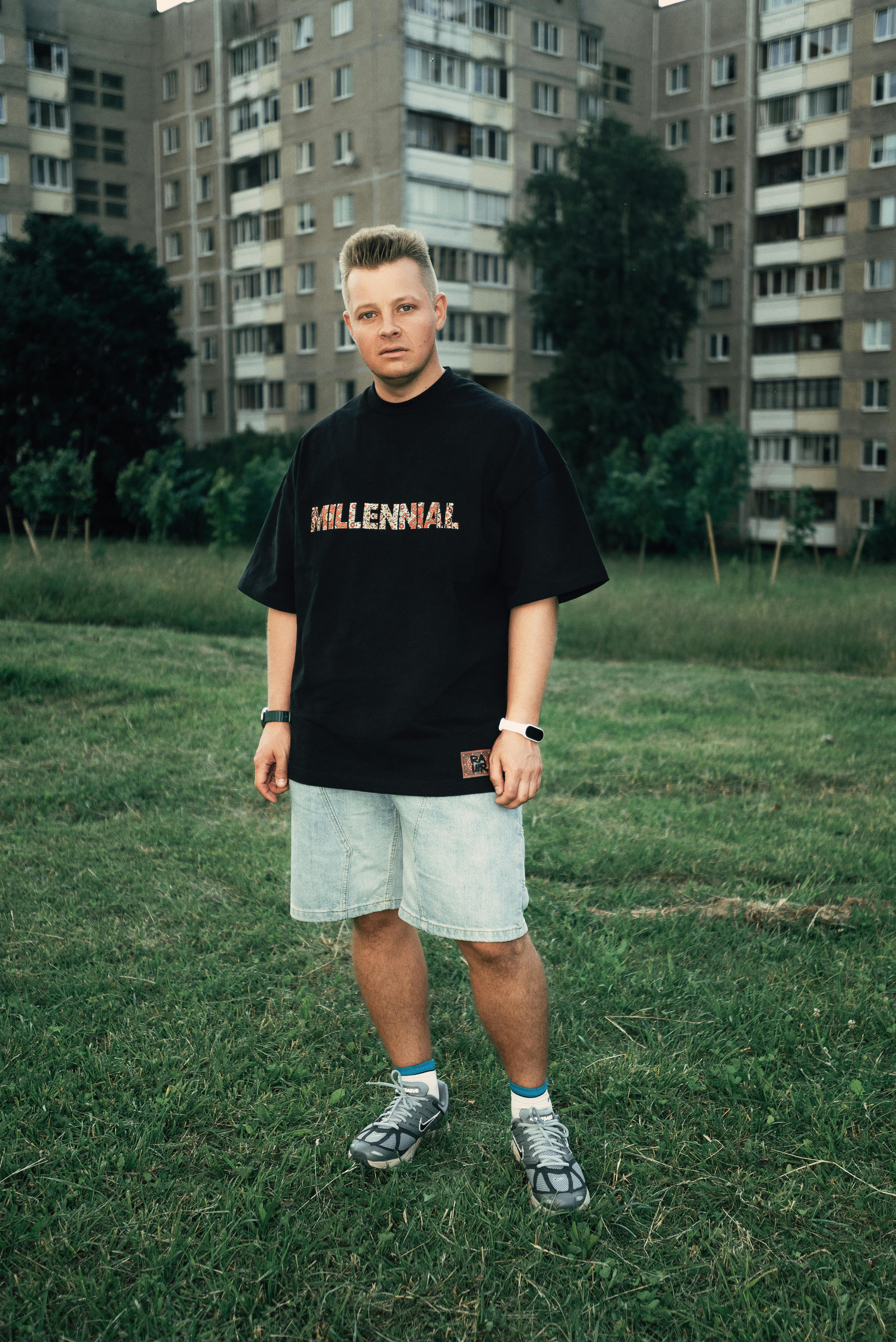 Casual portrait of a young man standing in a green urban park with apartment buildings in the background.