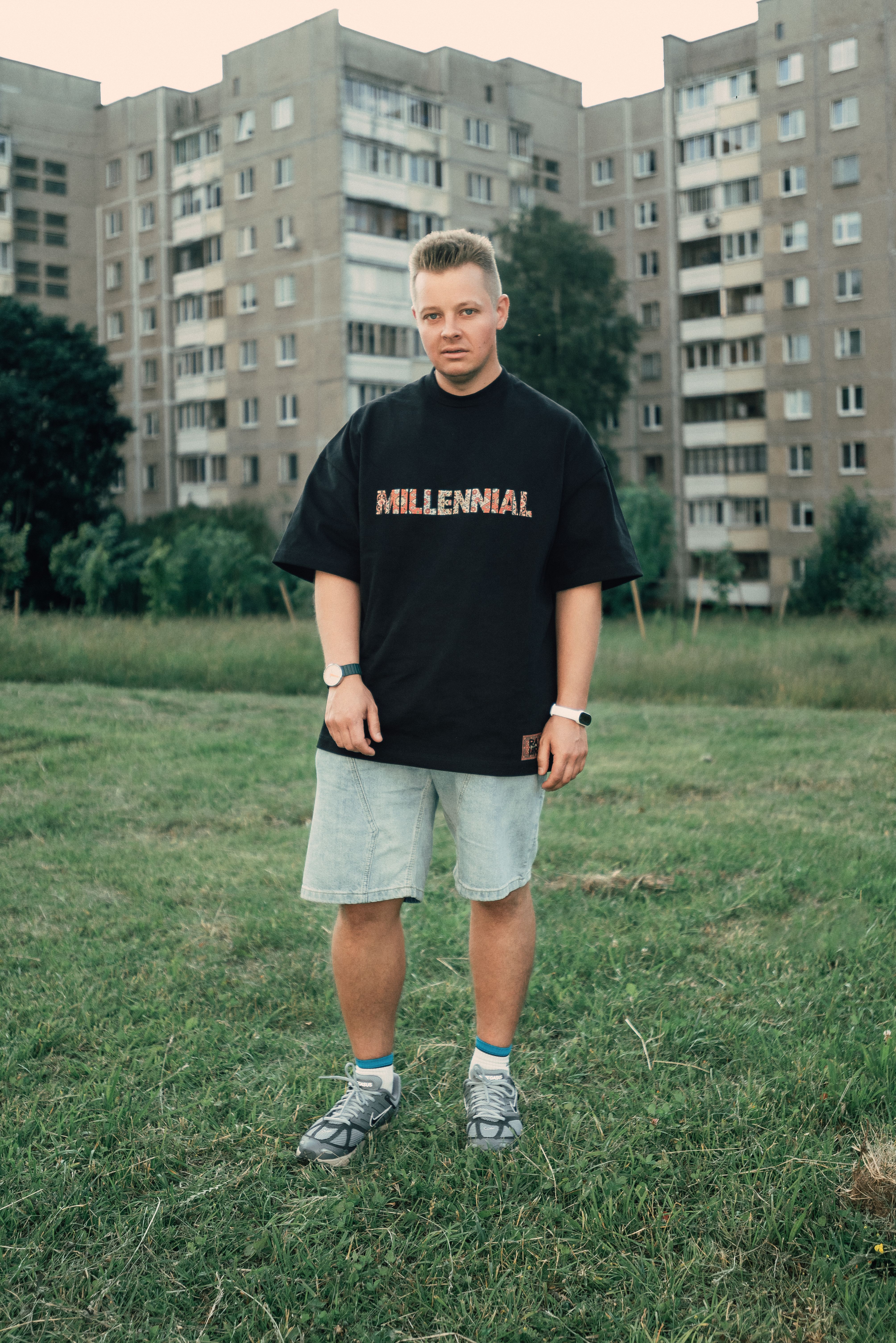 Young man stands casually in a grassy area with apartments in the background.