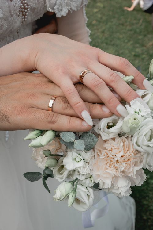 Free Close-up of intertwined hands with wedding rings holding an elegant floral bouquet. Stock Photo