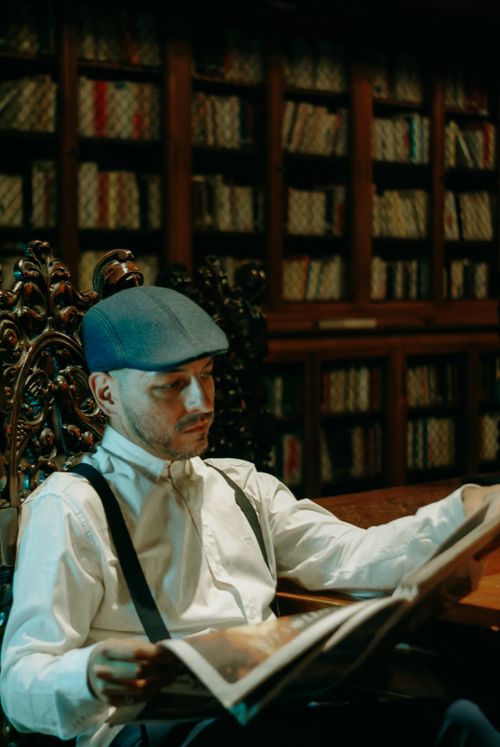 Free Adult man in a vintage library reading a newspaper, wearing suspenders and a flat cap. Stock Photo