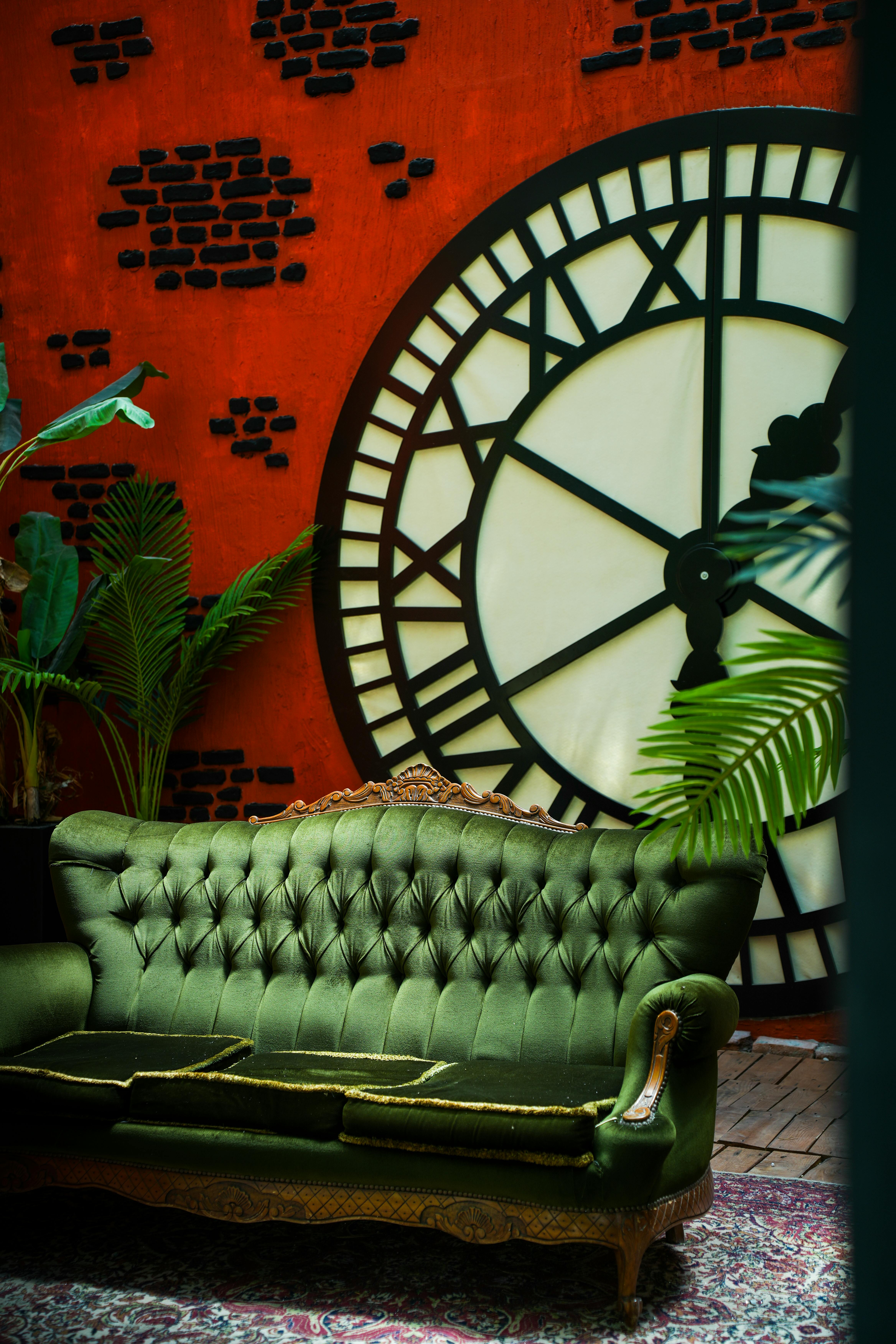 Elegant vintage sofa in front of large clock on rustic orange wall with lush green plants.
