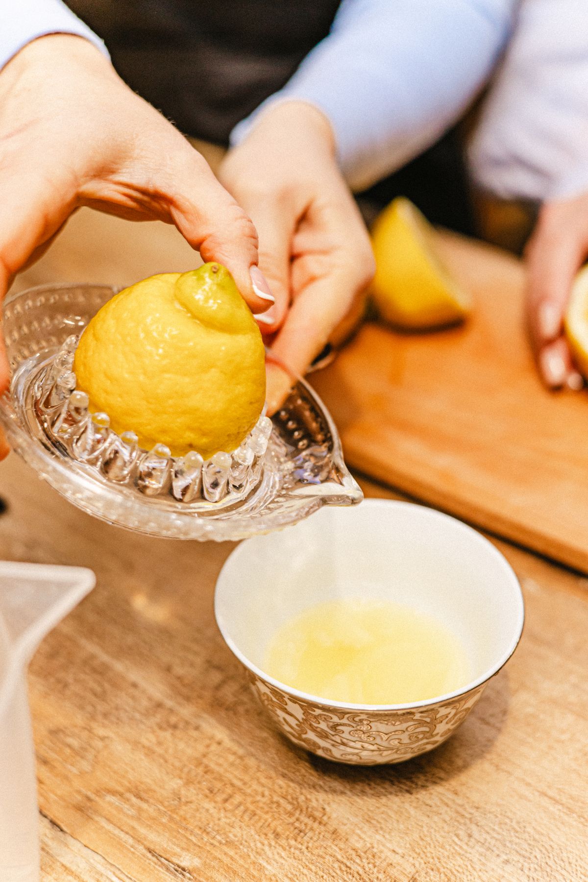 Hands pressing a lemon on a glass citrus juicer beside a small bowl